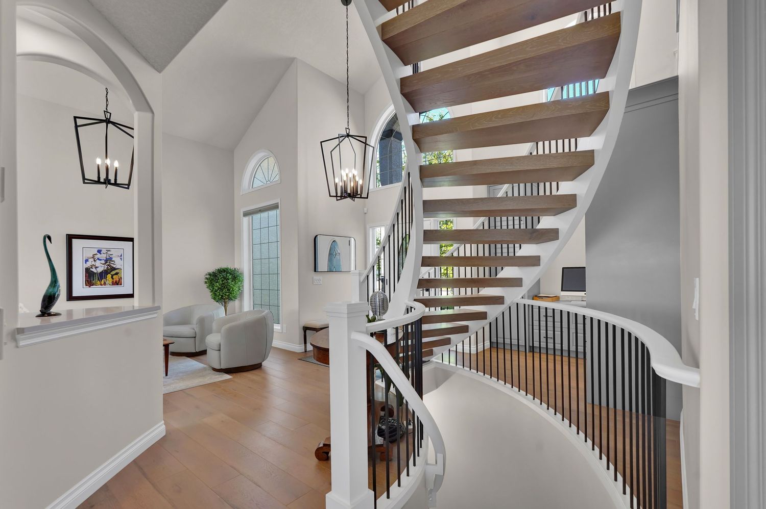 Elegant curved staircase with white balusters and wood steps in a bright foyer with arched windows and chandelier.