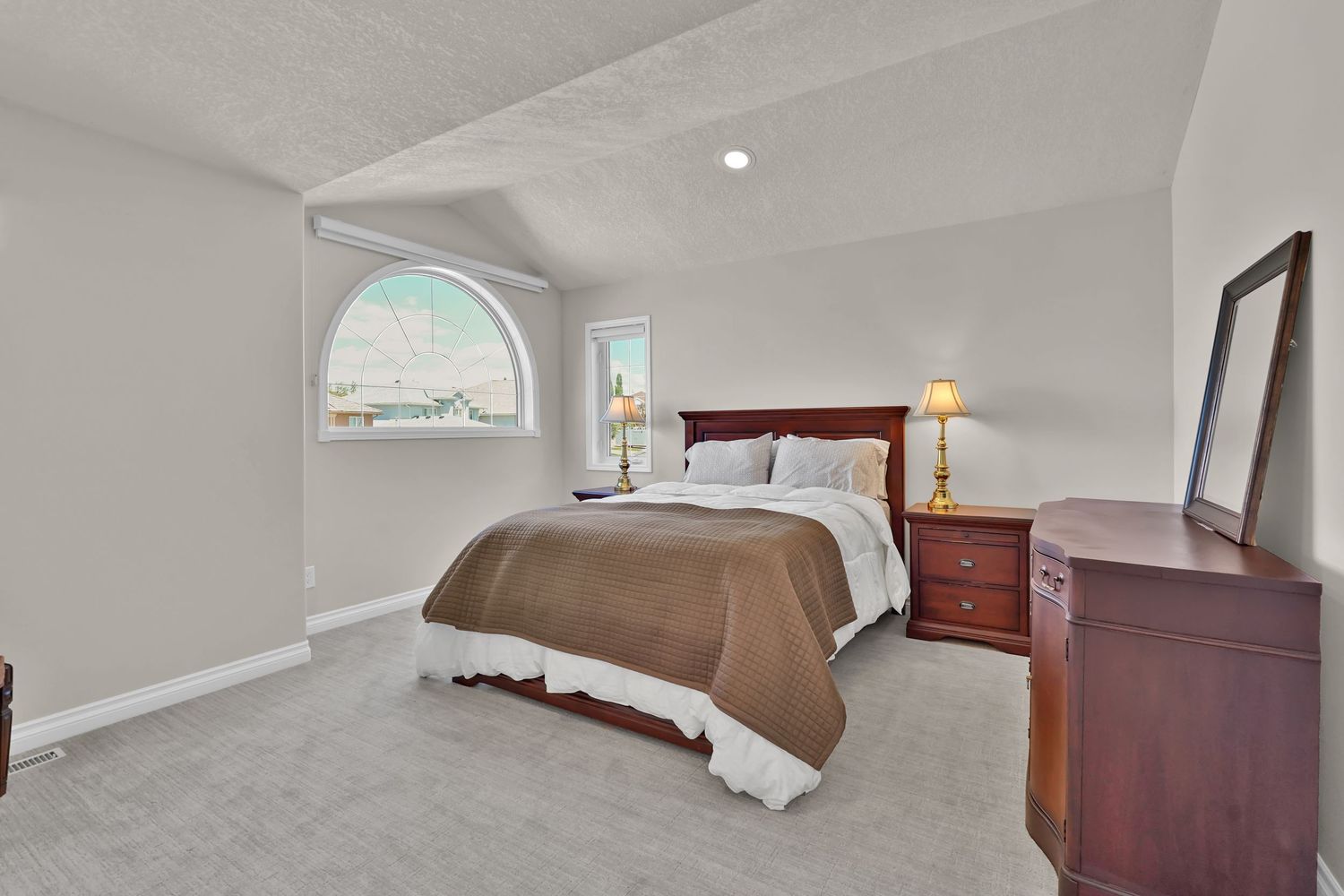 Spacious bedroom with vaulted ceiling, arched window, plush carpet, wooden dresser with mirror, and neutral decor.