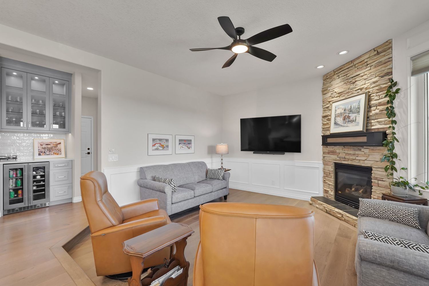 Cozy living room with stone fireplace, ceiling fan, wall-mounted TV, leather chairs, and built-in bar cabinet.
