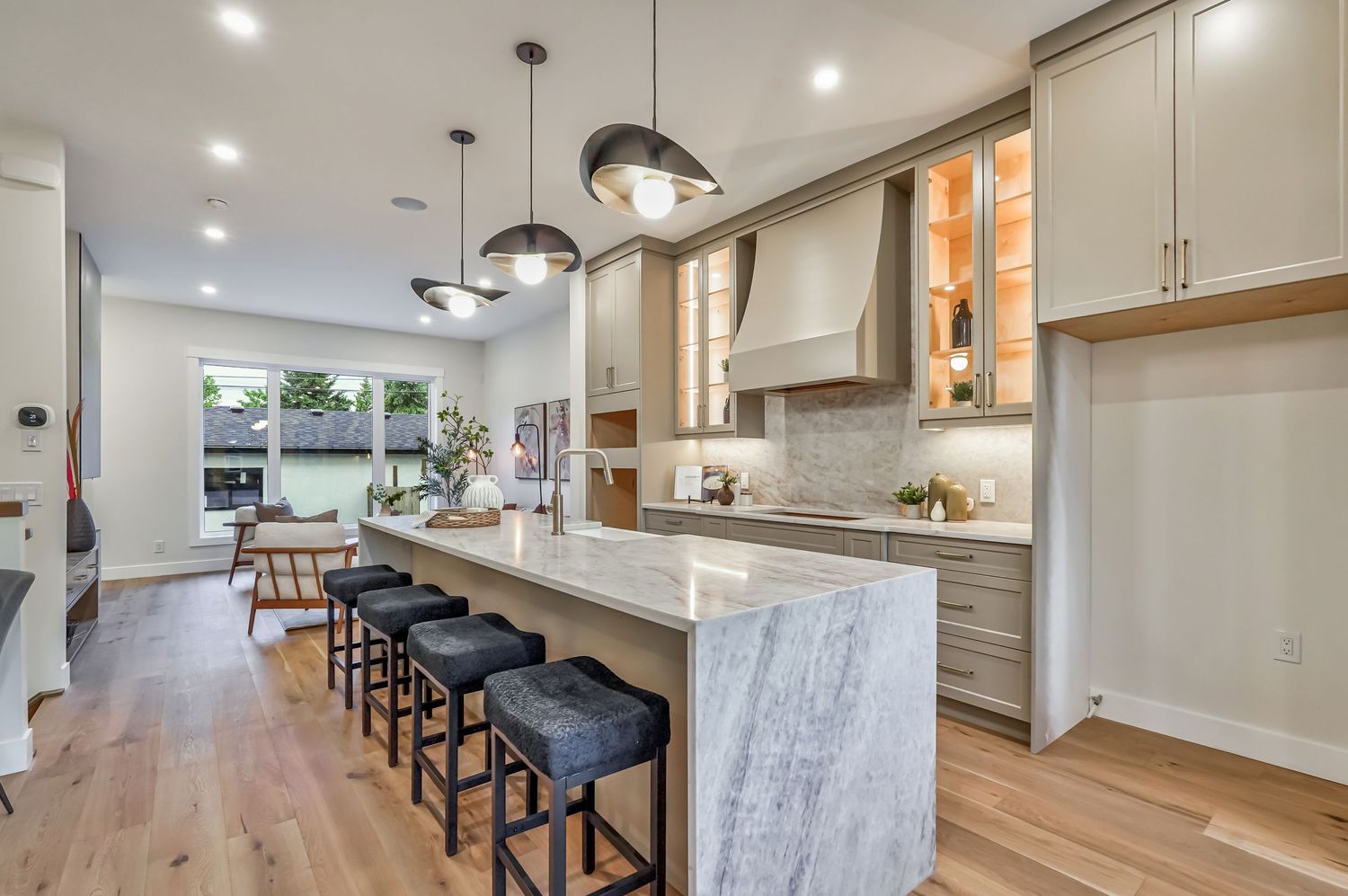 Modern kitchen with marble island, pendant lights, gray cabinets, bar stools, and hardwood floors in an open-plan home.