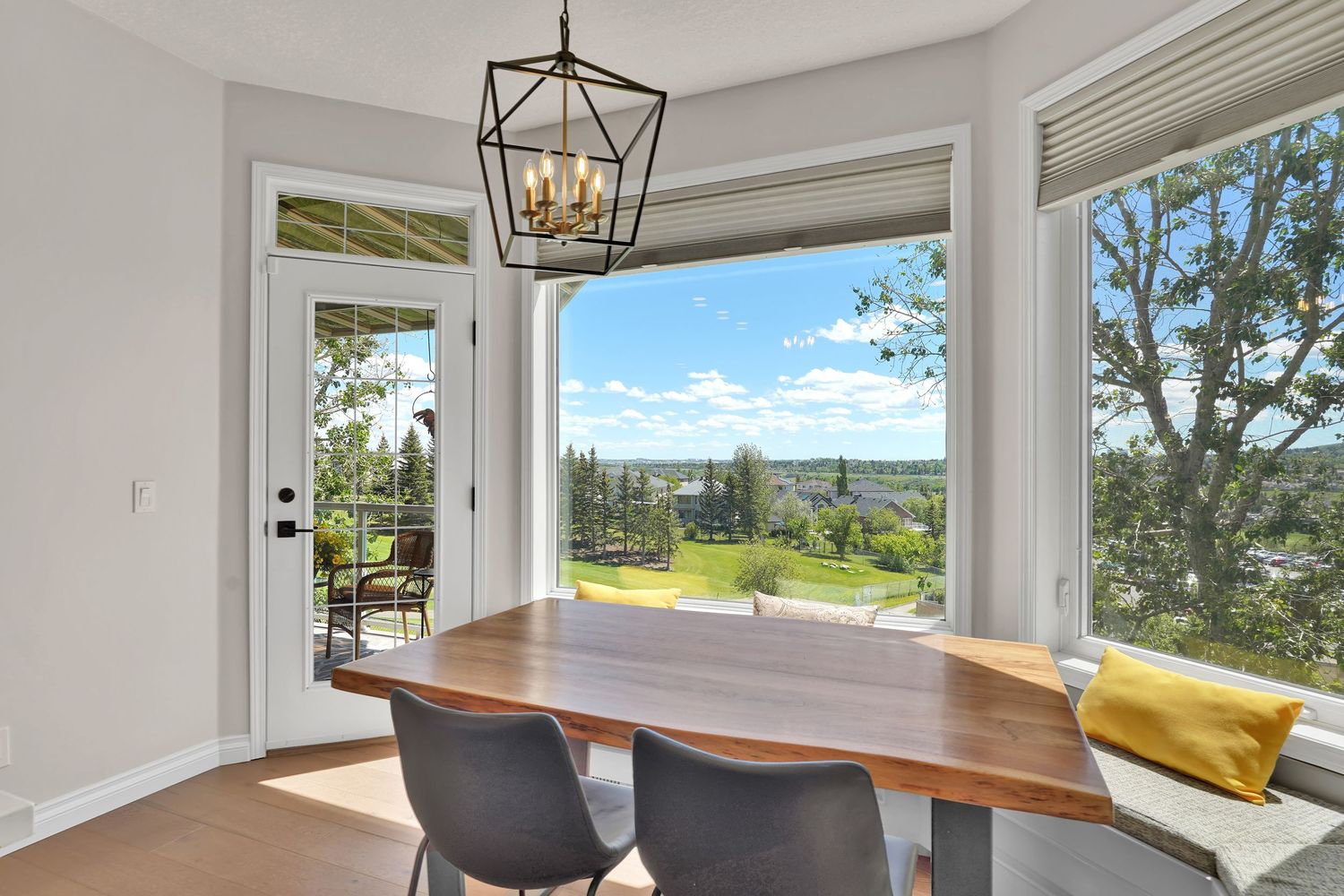Bright dining area with wood table, modern chandelier, and large windows offering panoramic views of lush greenery.