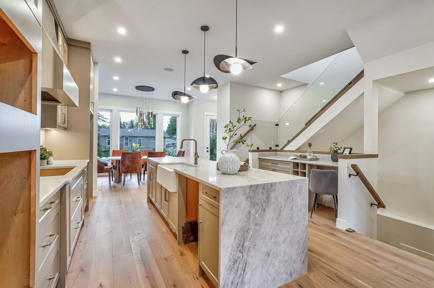 Modern open-concept kitchen with marble island, pendant lights, light wood floors, and natural wood cabinetry leading to dining area.
