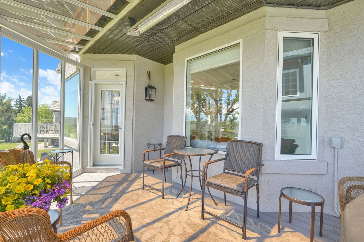 Covered patio with bistro table, chairs, and wicker seating surrounded by bright windows and colorful flowers.