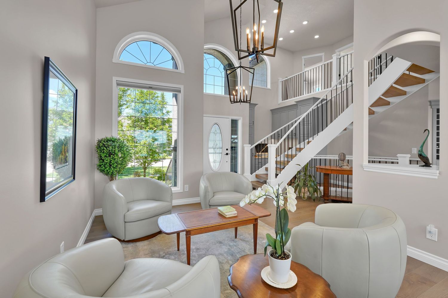 Bright two-story living room with vaulted ceilings, chandelier, white furniture, wood coffee table, and elegant staircase.