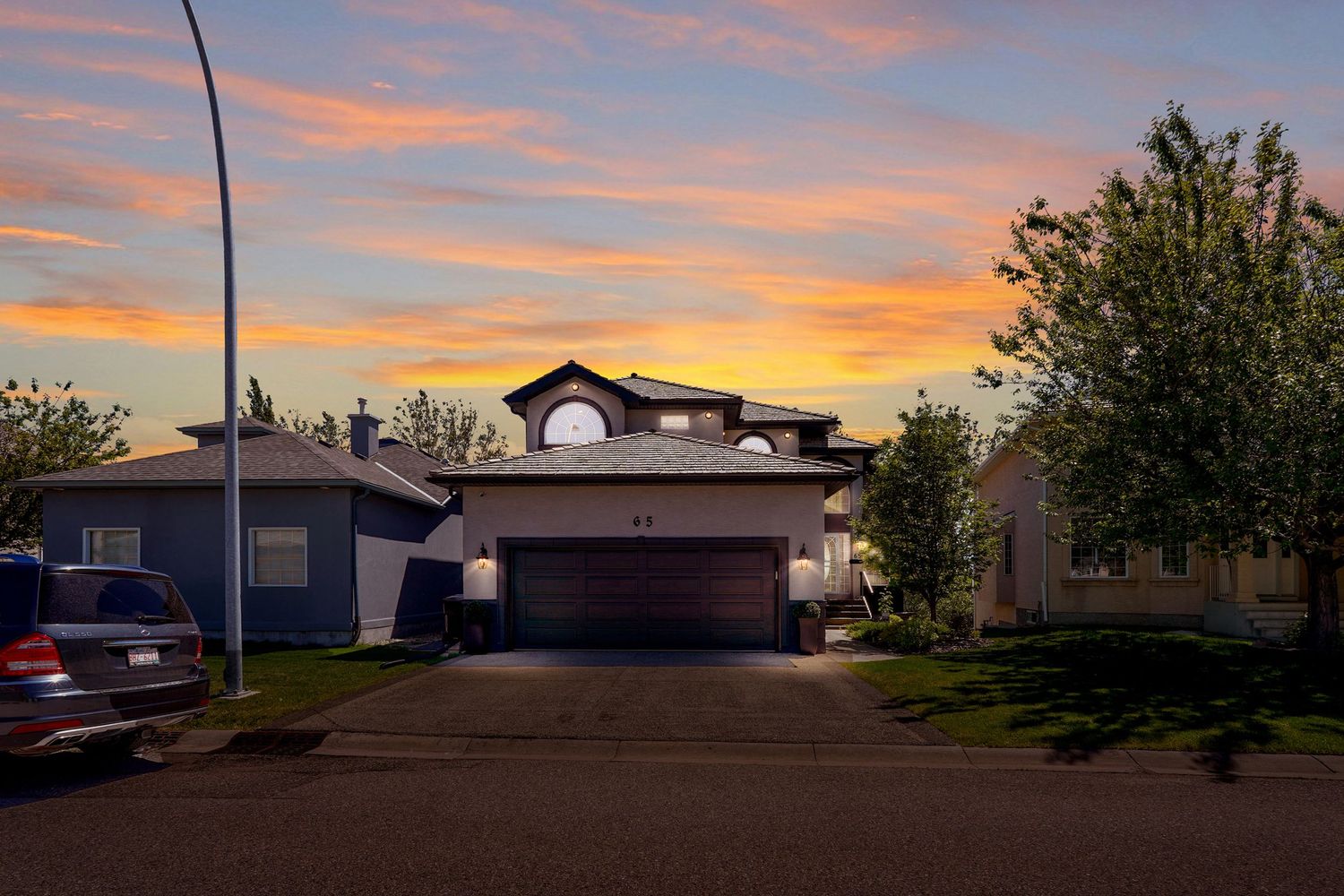 Suburban two-story home with garage at sunset, vibrant orange and purple sky, green trees lining quiet residential street.