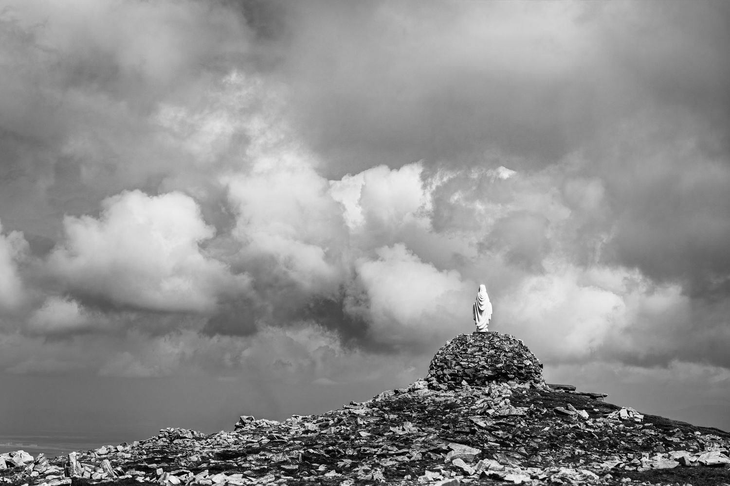 Black and white photo of a white statue atop a rocky hill under dramatic cloudy sky.