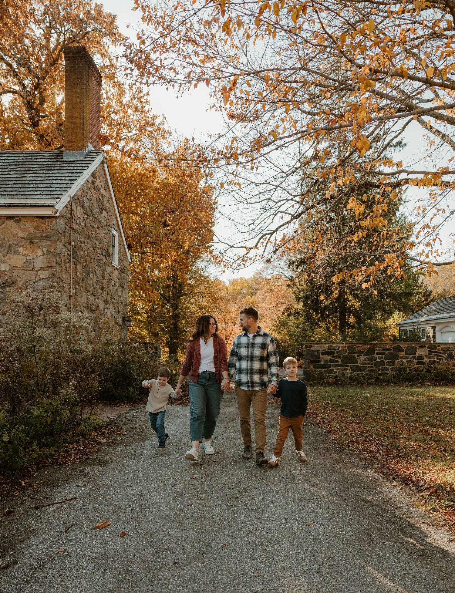 Family of four walking hand-in-hand down a scenic autumn path lined with stone buildings and colorful fall foliage.