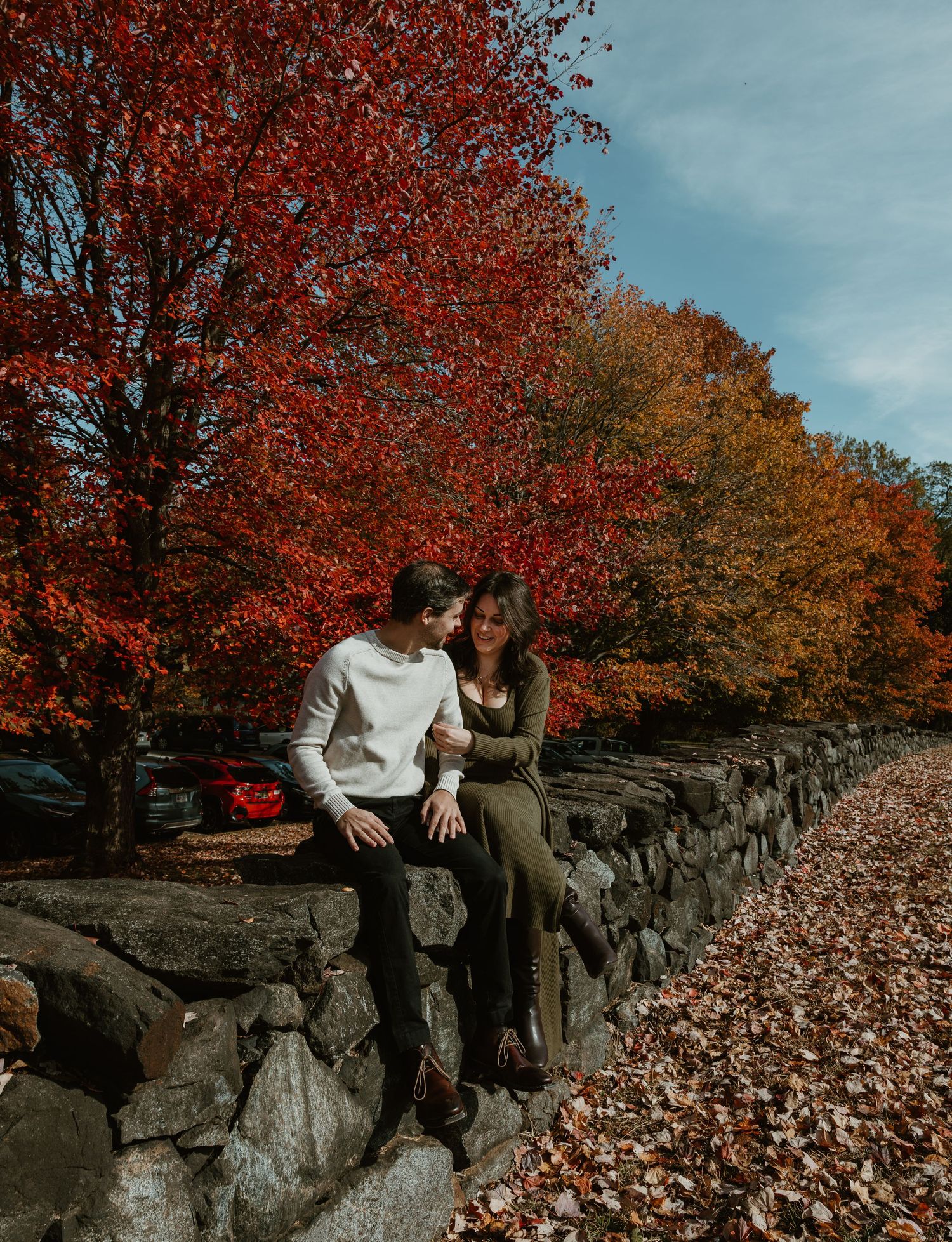 Couple sitting on a stone wall surrounded by vibrant red and orange autumn trees on a sunny fall day.