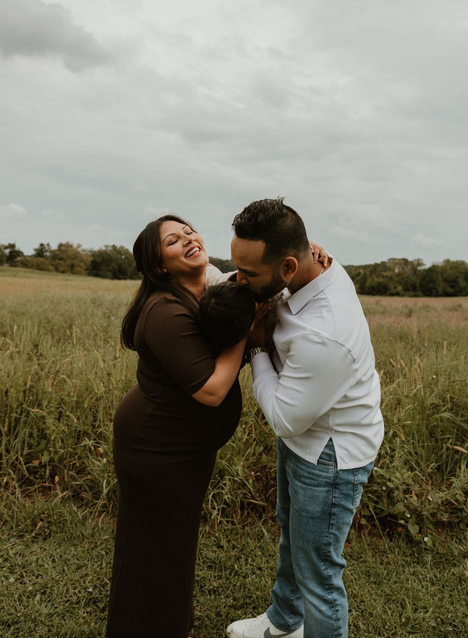 Couple laughing and embracing in a grassy field, woman in brown dress, man in white shirt and jeans, cloudy sky.