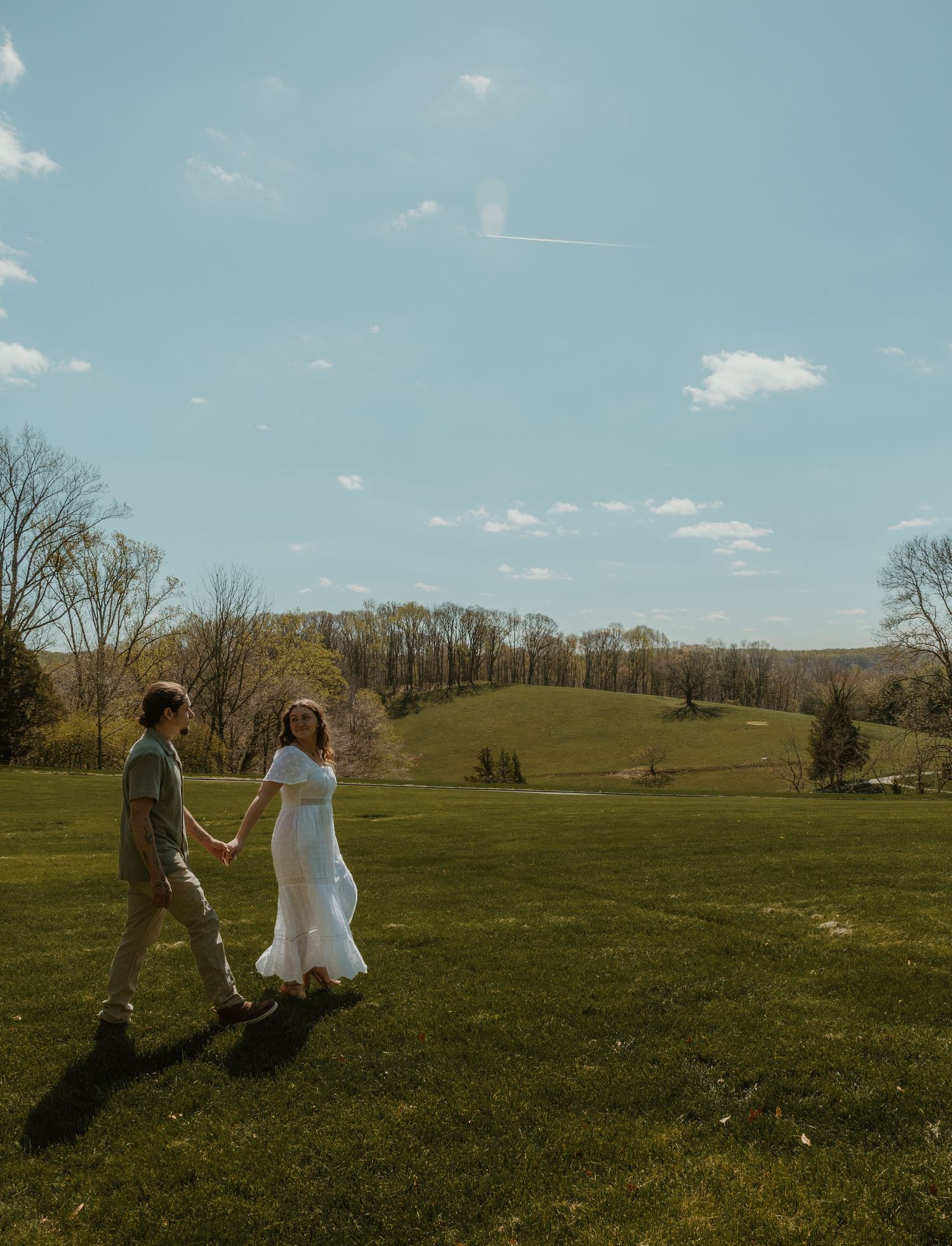 Couple holding hands walking across green meadow in white dress and gray suit under blue sky.