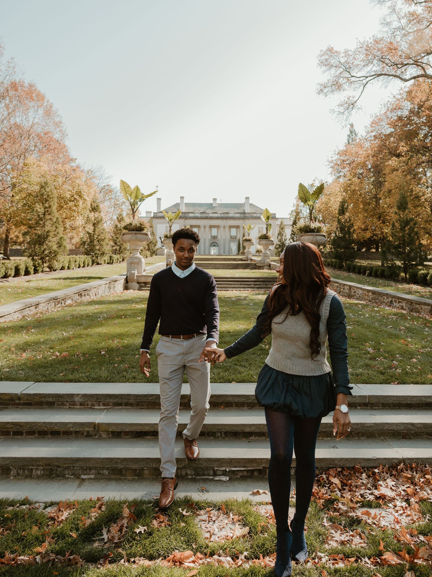 Couple holding hands walking up stone steps in an autumn garden with a historic mansion in the background.