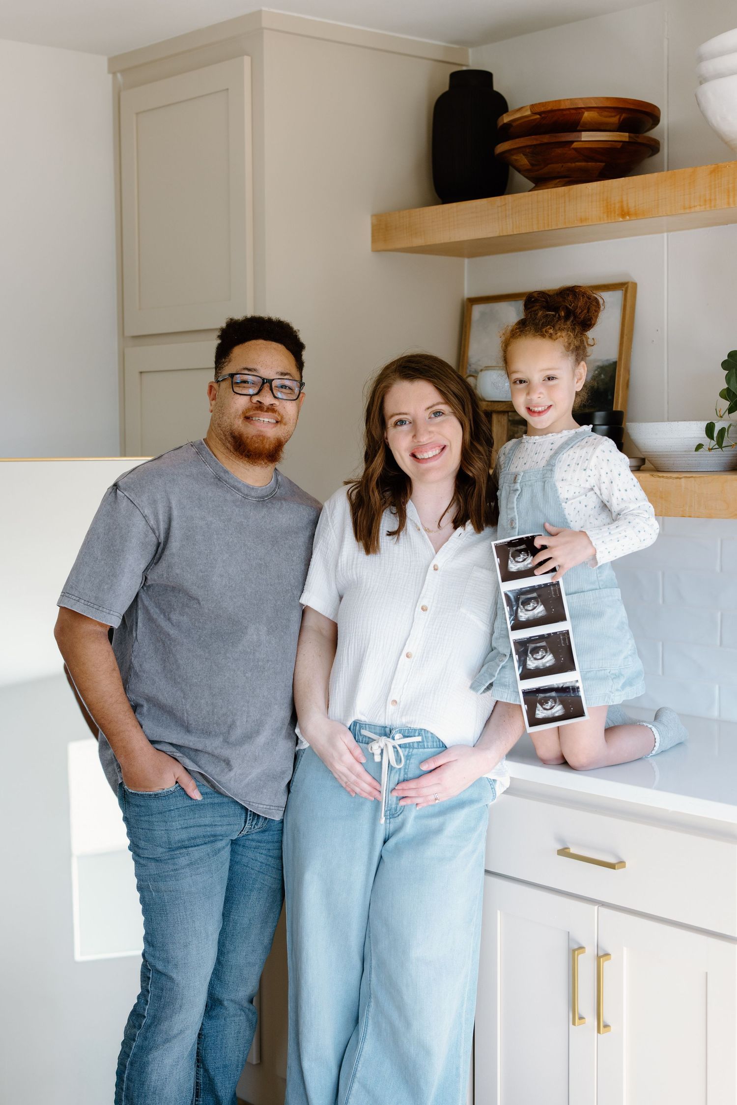 Happy family pregnancy announcement with couple and young girl holding ultrasound photos in a bright kitchen.