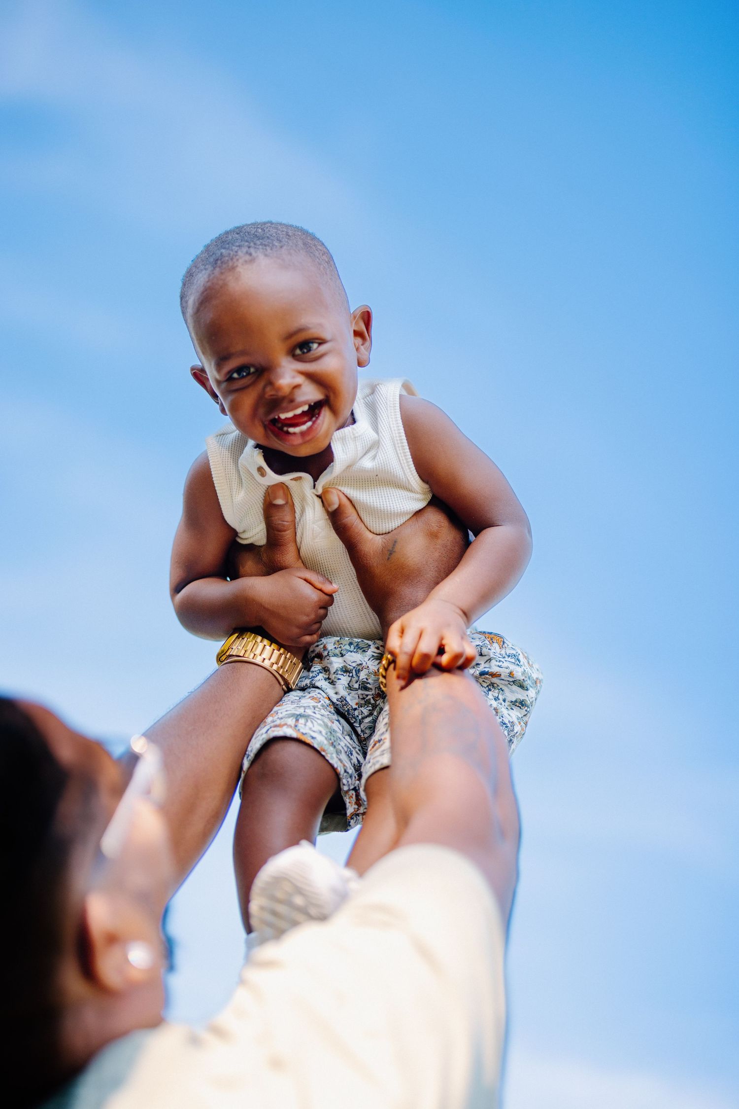 A joyful child in a white tank top and patterned shorts is lifted up against a bright blue sky.