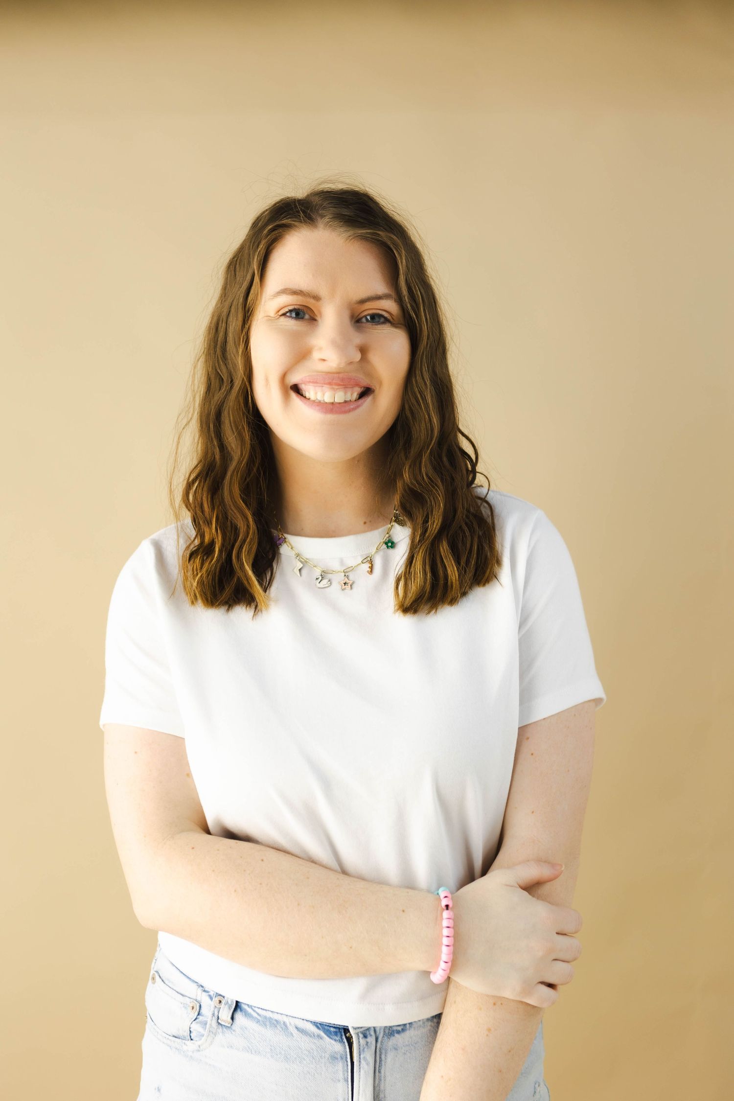 A person with brunette wavy hair wearing a white t-shirt and jeans poses against a beige background with a cheerful expression.