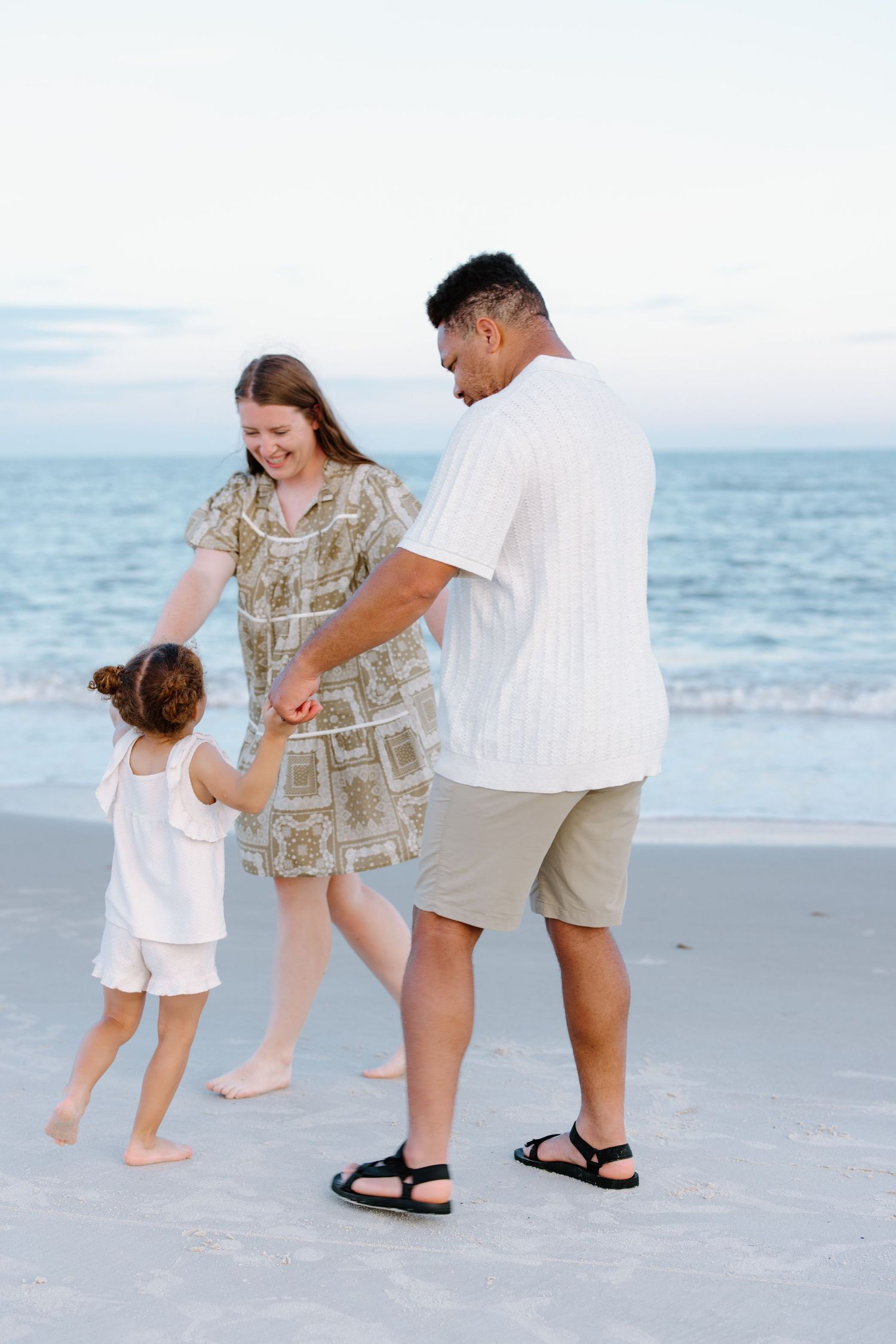 Family of three enjoying a beach walk at sunset, parents holding hands with young child near ocean waves.