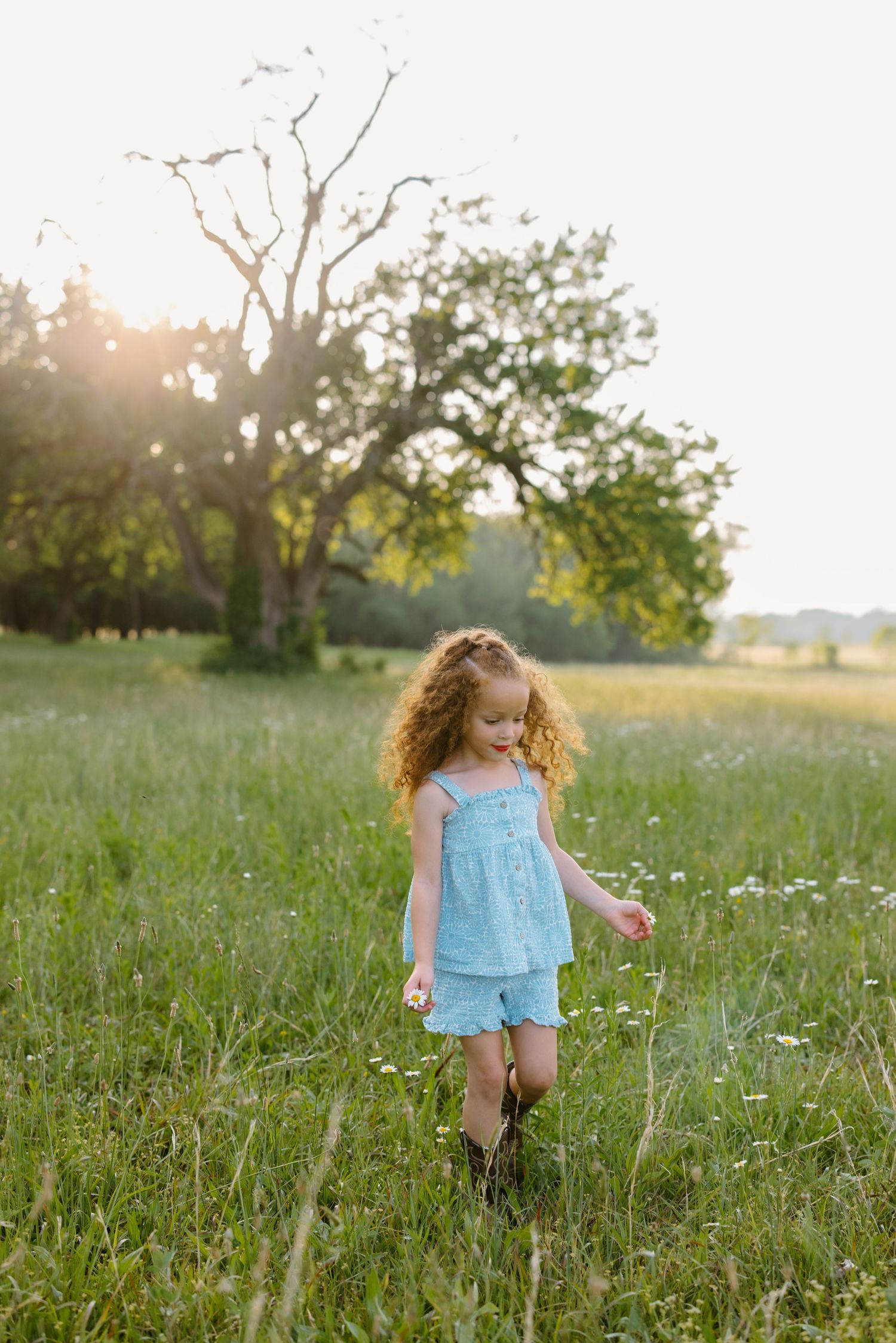 A curly-haired girl in a blue dress walks through a sunny green meadow with wildflowers and trees in the background.