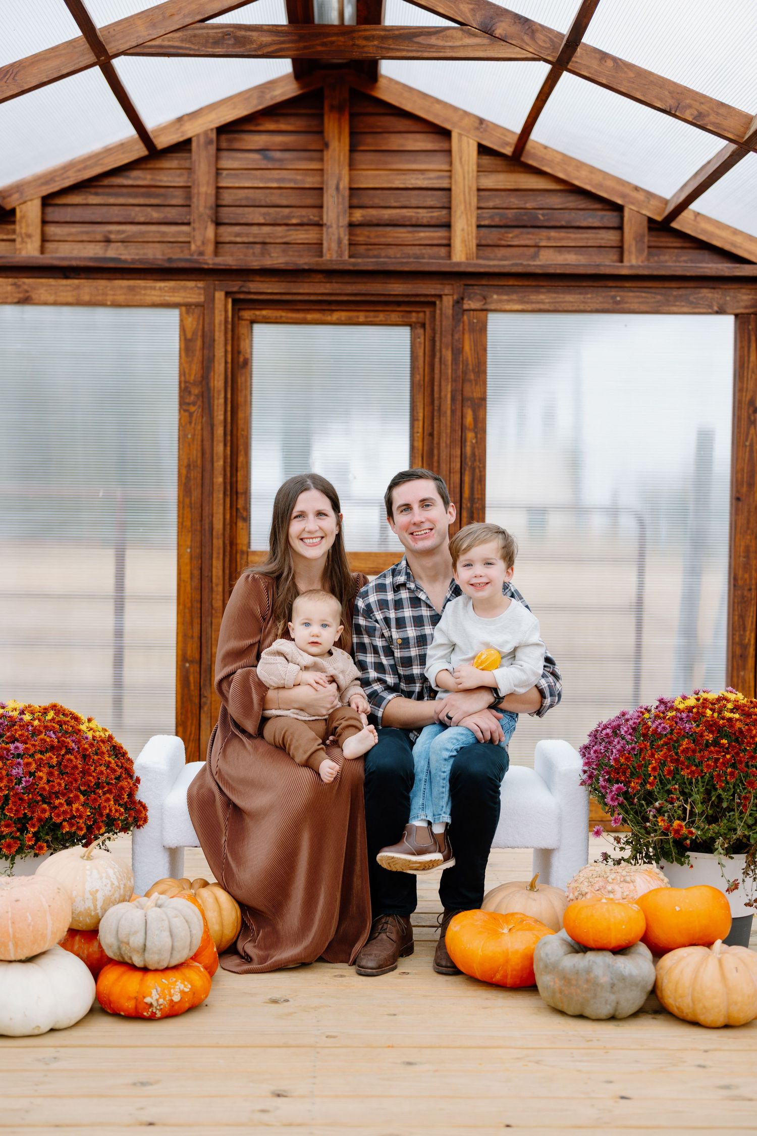 A cozy greenhouse decorated for fall with orange pumpkins and red mums by the entrance on a wooden deck.