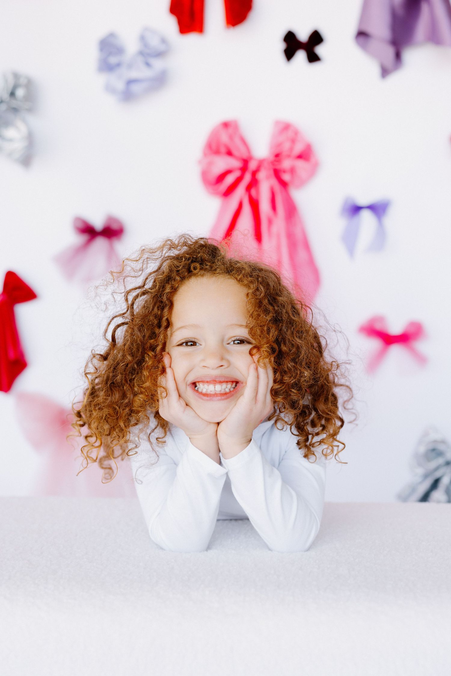 A child with curly red hair in a white shirt poses against a background decorated with colorful bows.