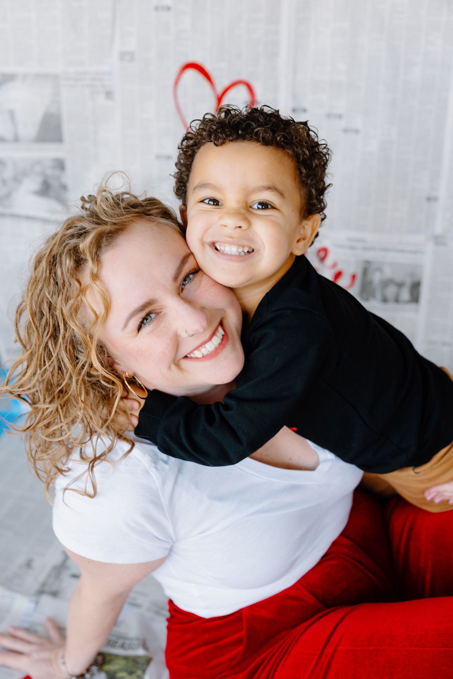 A sweet portrait of parent and child sharing hugs and smiles against a white wall with red hearts.
