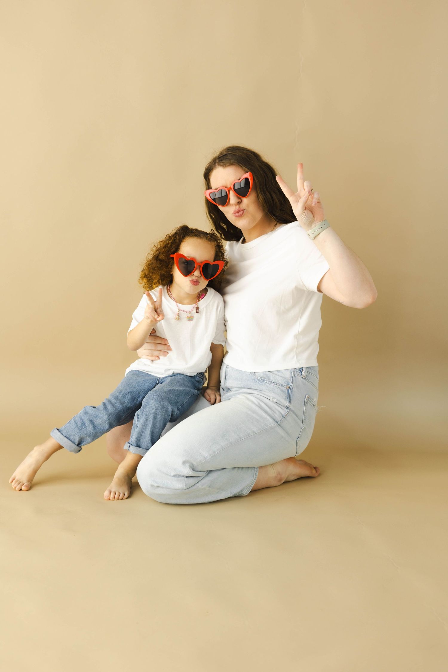 Mother and daughter in matching white tees, jeans, and red heart sunglasses posing with peace signs on beige background.