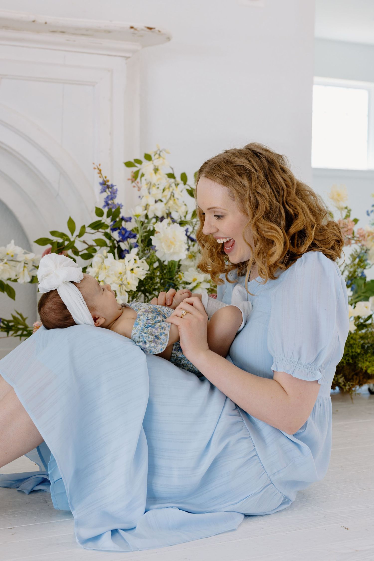 A tender moment with woman in light blue dress holding a newborn baby surrounded by floral arrangements.