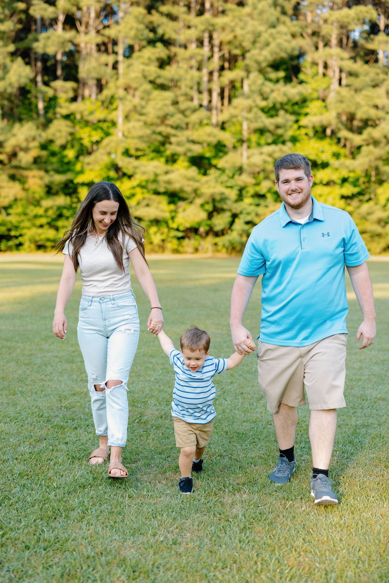Family walking together and holding hands on grass field with pine forest in background.