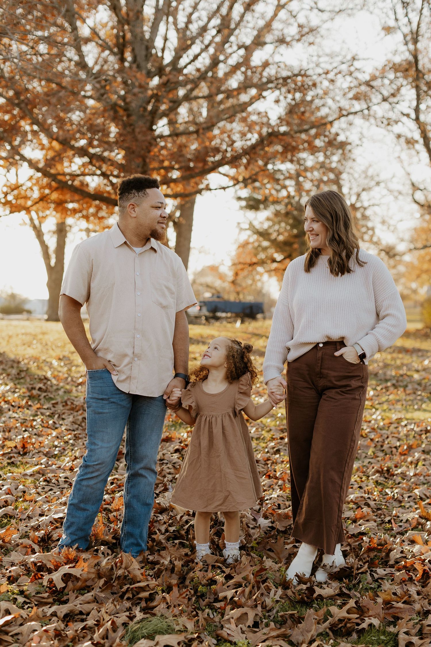 Family of three holding hands in autumn park, parents smiling at toddler girl in brown dress among fallen leaves.
