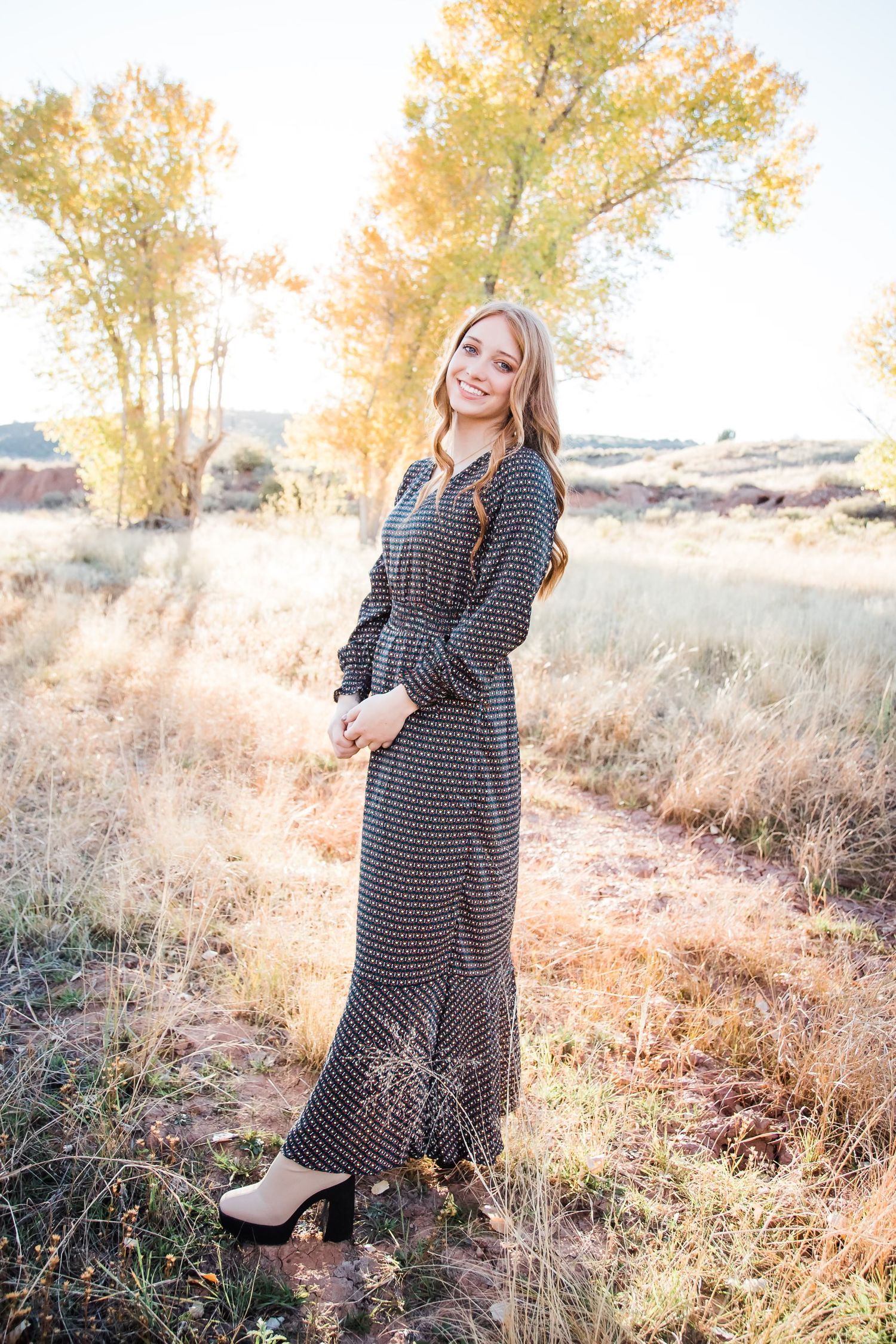 A person poses in a flowing maxi dress against autumn trees during golden hour.