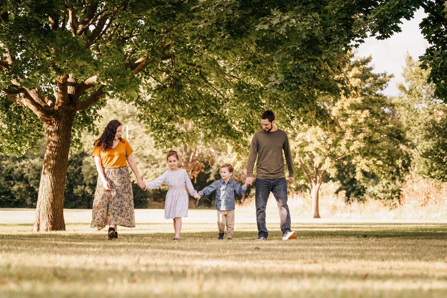 A family walks together holding hands under tree branches in a sunlit park.