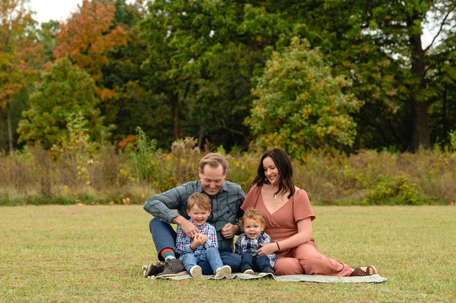 A family sits together on a blanket in an autumn field with fall foliage in the background.