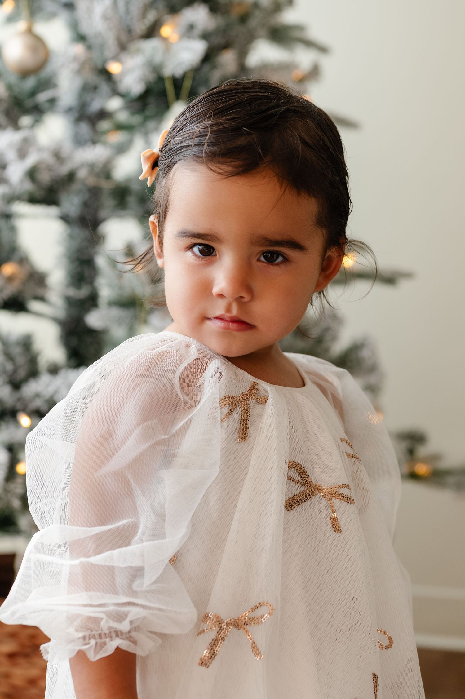 Child wearing white ruffled dress with gold bows poses by Christmas tree.