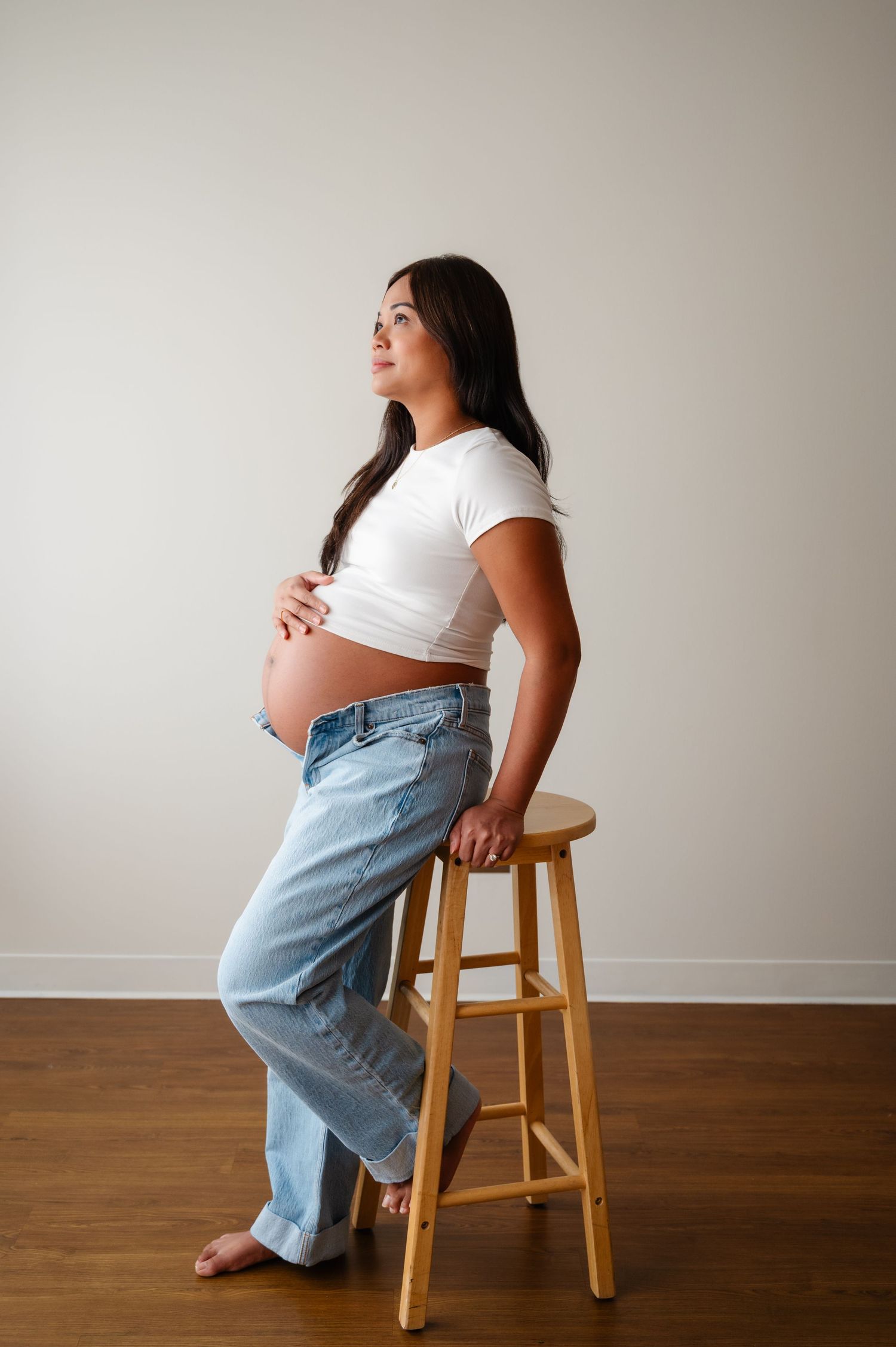A pregnant woman in white shirt and jeans sits on a wooden stool against a neutral wall in a maternity photo series.