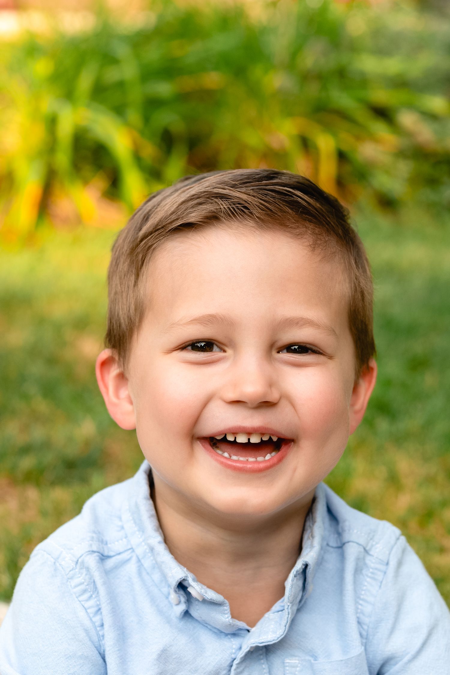 Young child in light blue shirt smiling brightly outdoors in natural setting.