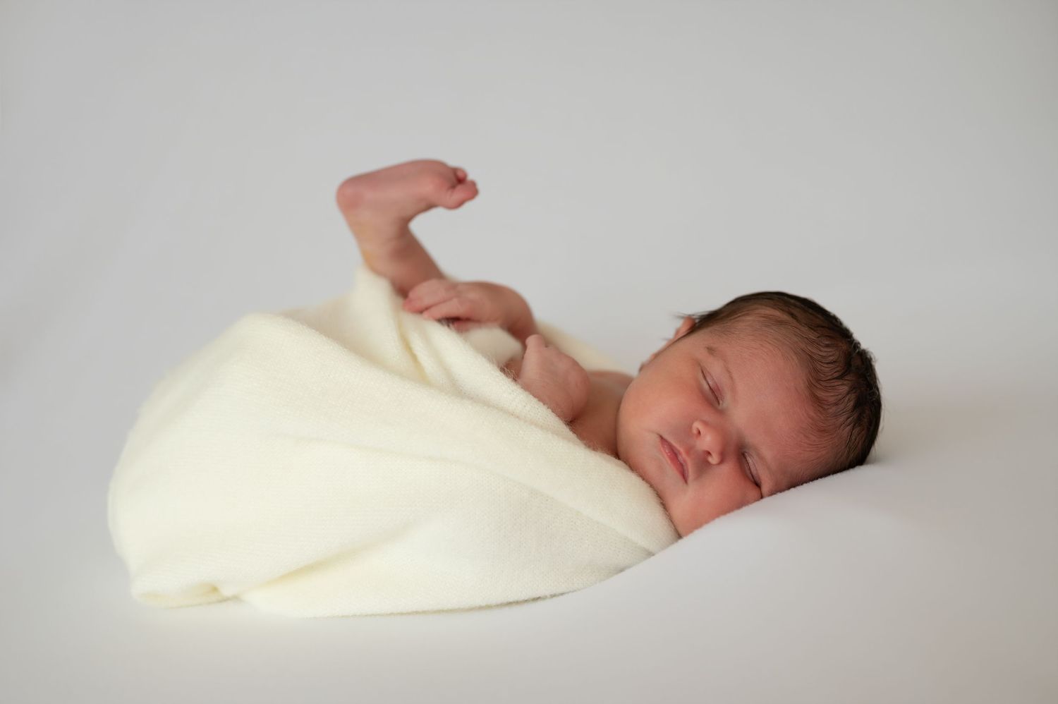 A peaceful newborn baby wrapped in a soft cream-colored blanket sleeps on a white background.