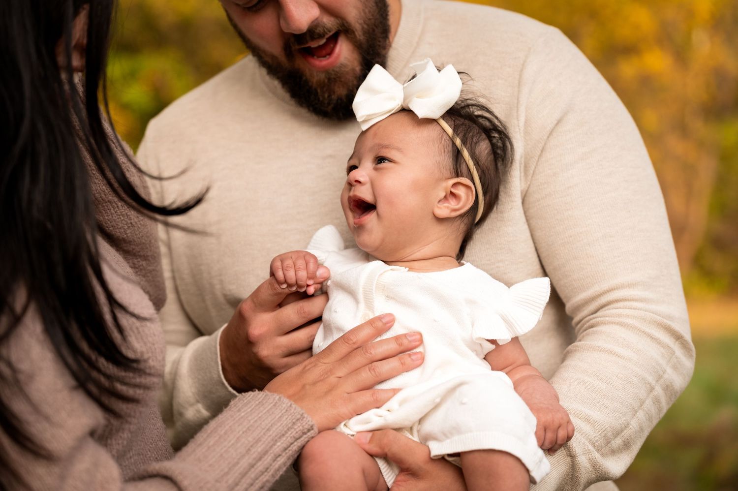 A laughing baby in white dress being held during an autumn outdoor photoshoot.