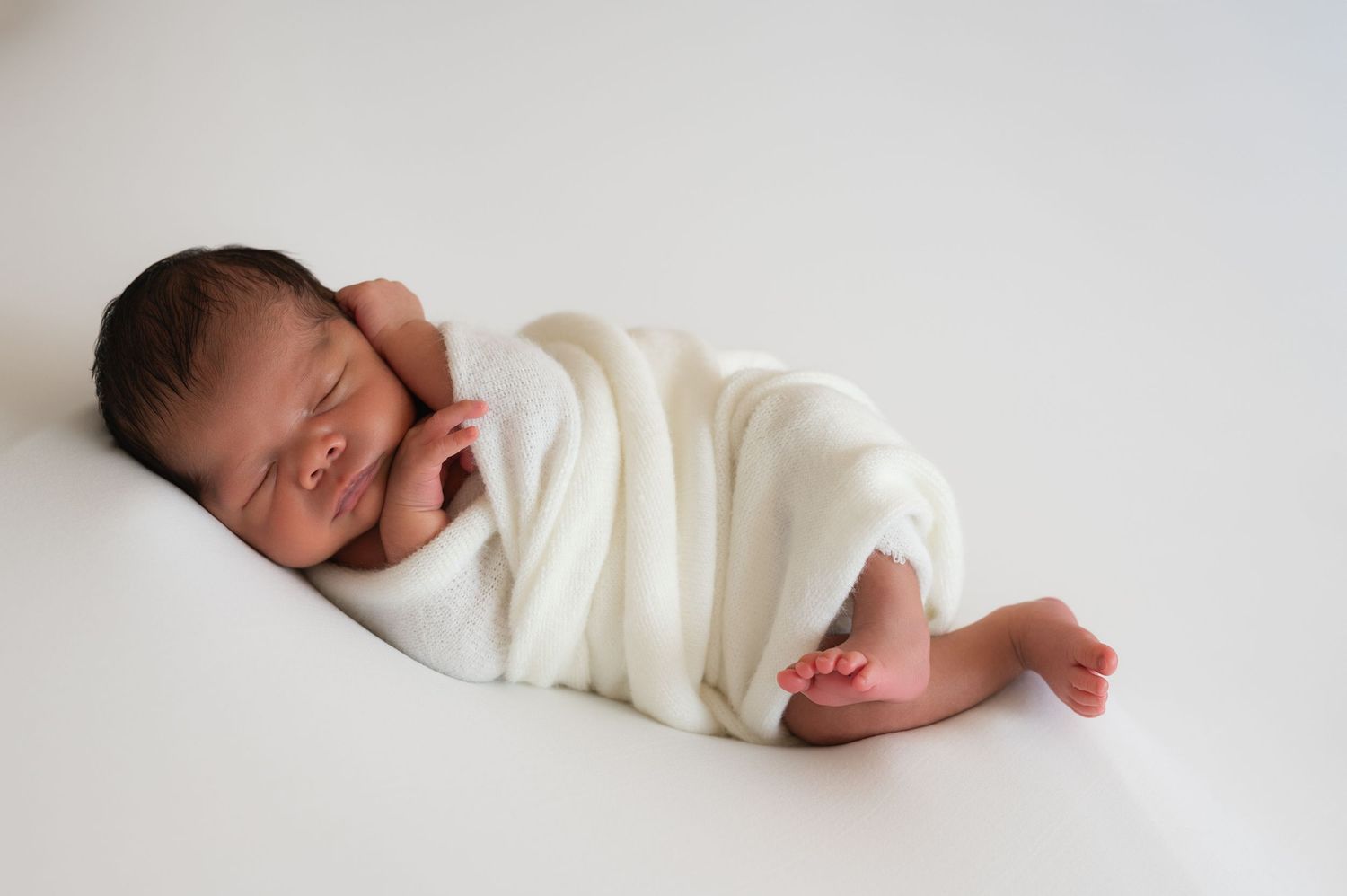 Newborn baby wrapped in white blanket sleeping peacefully on white background.