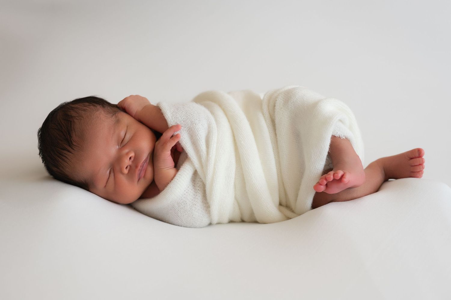 Sleeping newborn baby wrapped in white blanket on minimalist white backdrop.