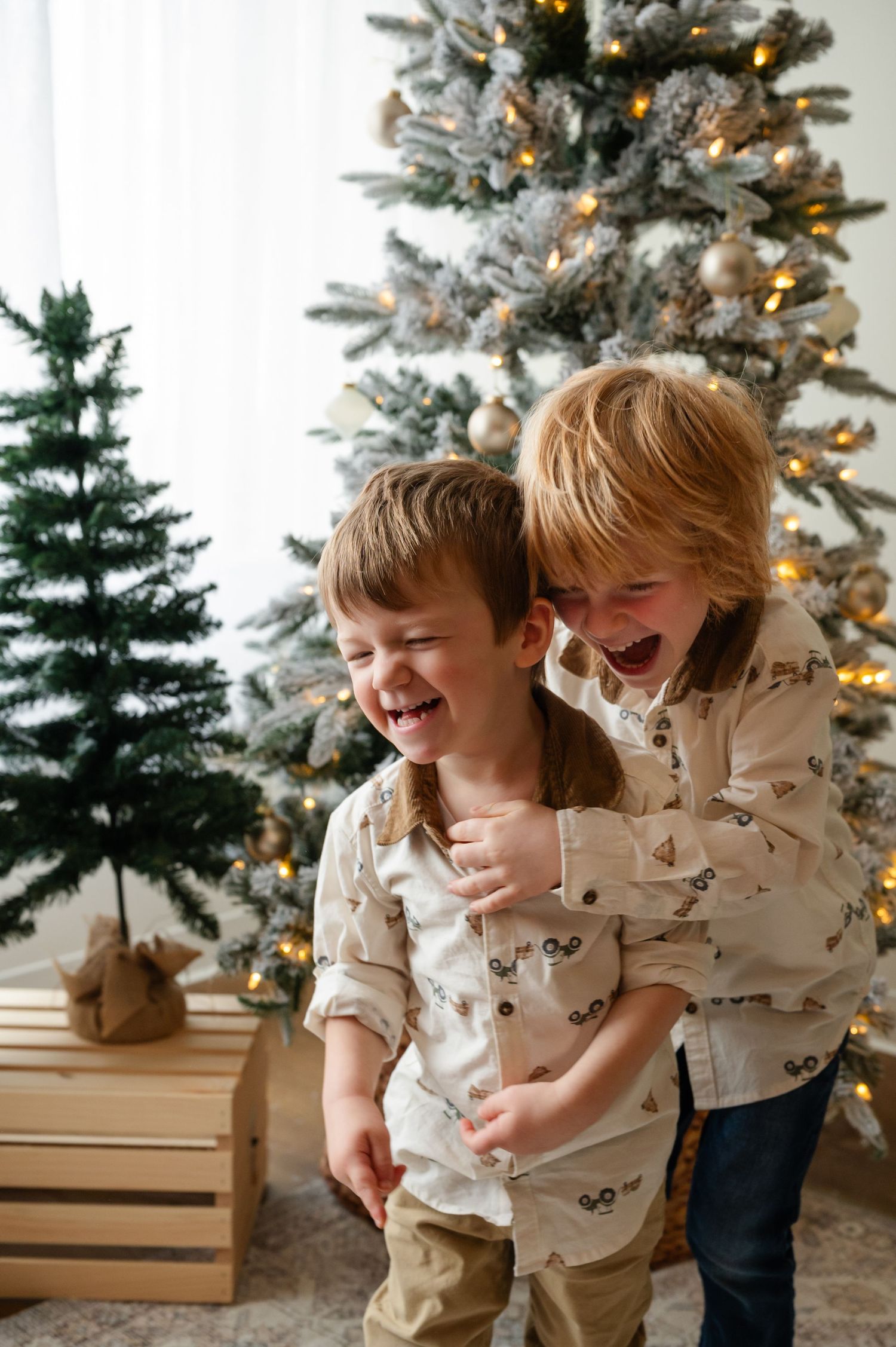 Two children in matching outfits laugh and play in front of lit Christmas tree.