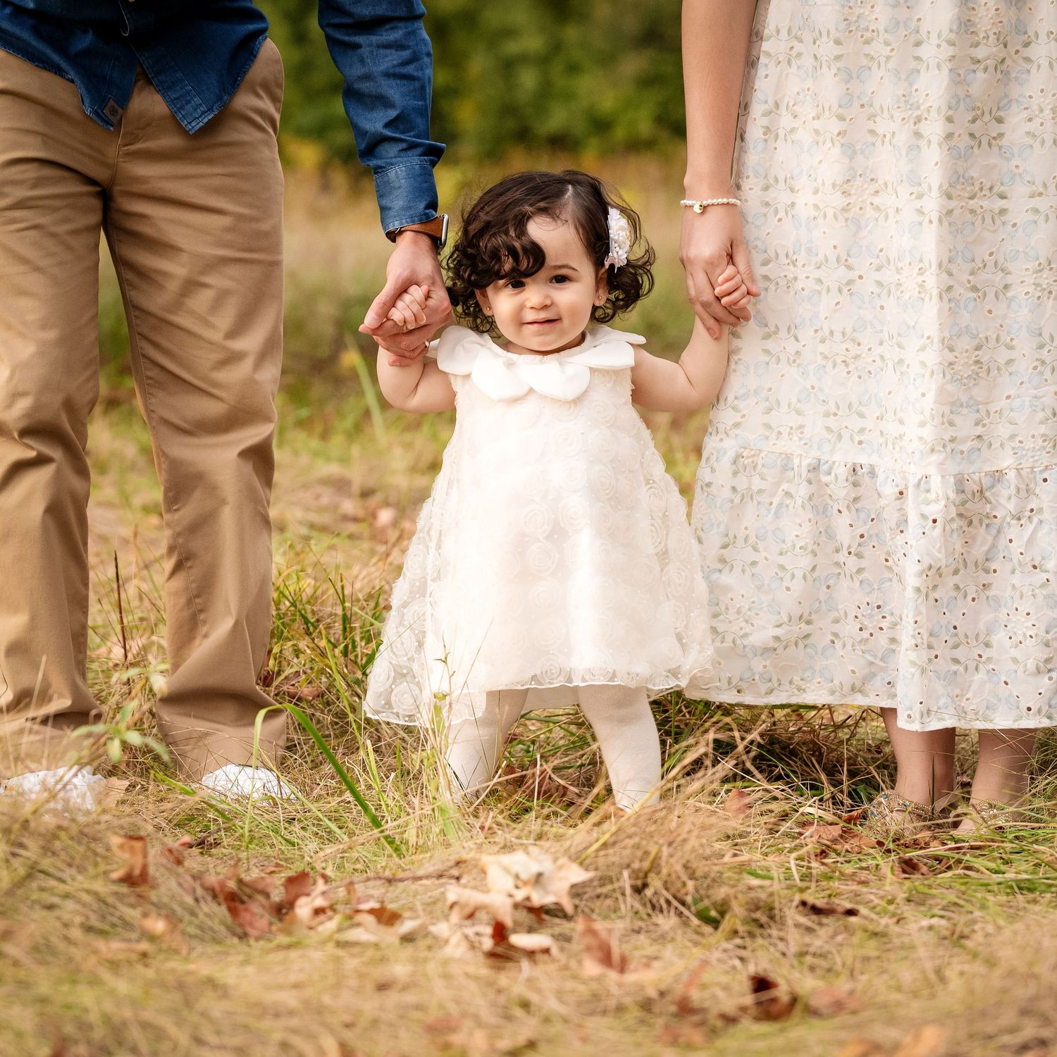 A small child in a white dress holds hands with parents while walking through autumn leaves.