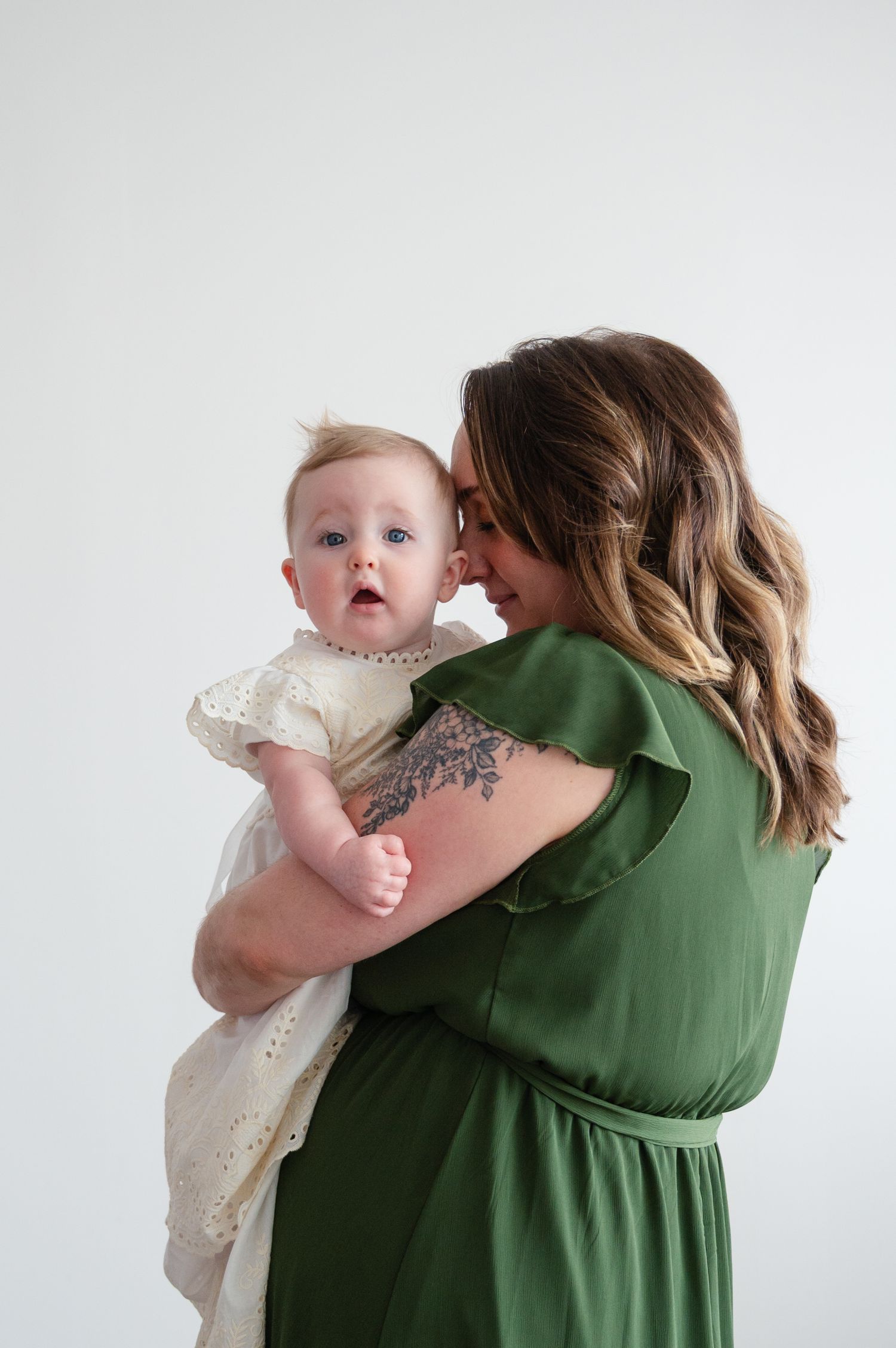 An intimate indoor portrait showing a figure in green holding a baby in a white lace dress.