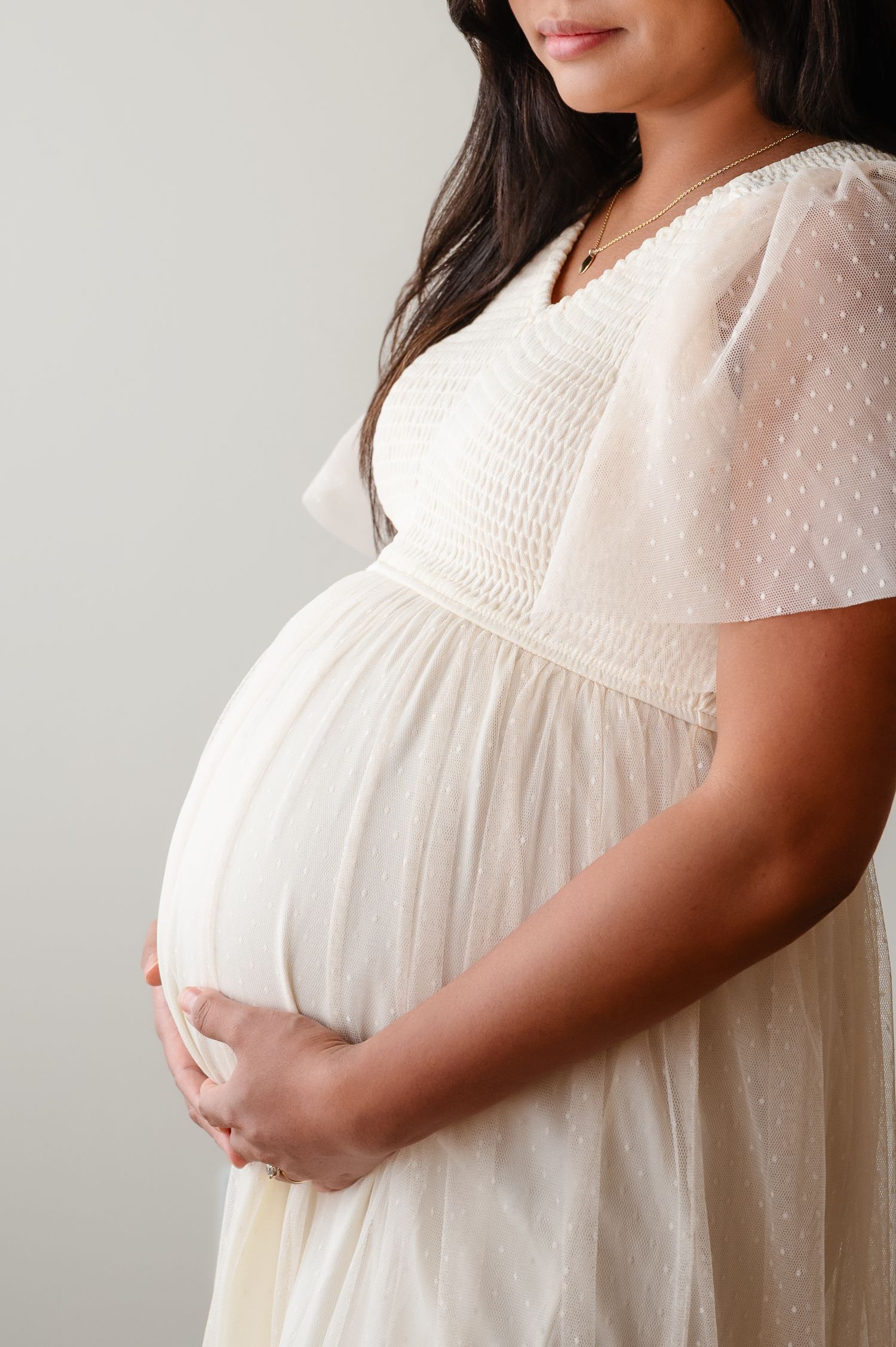 Expectant mother in white lace dress cradles her baby bump against a light background.