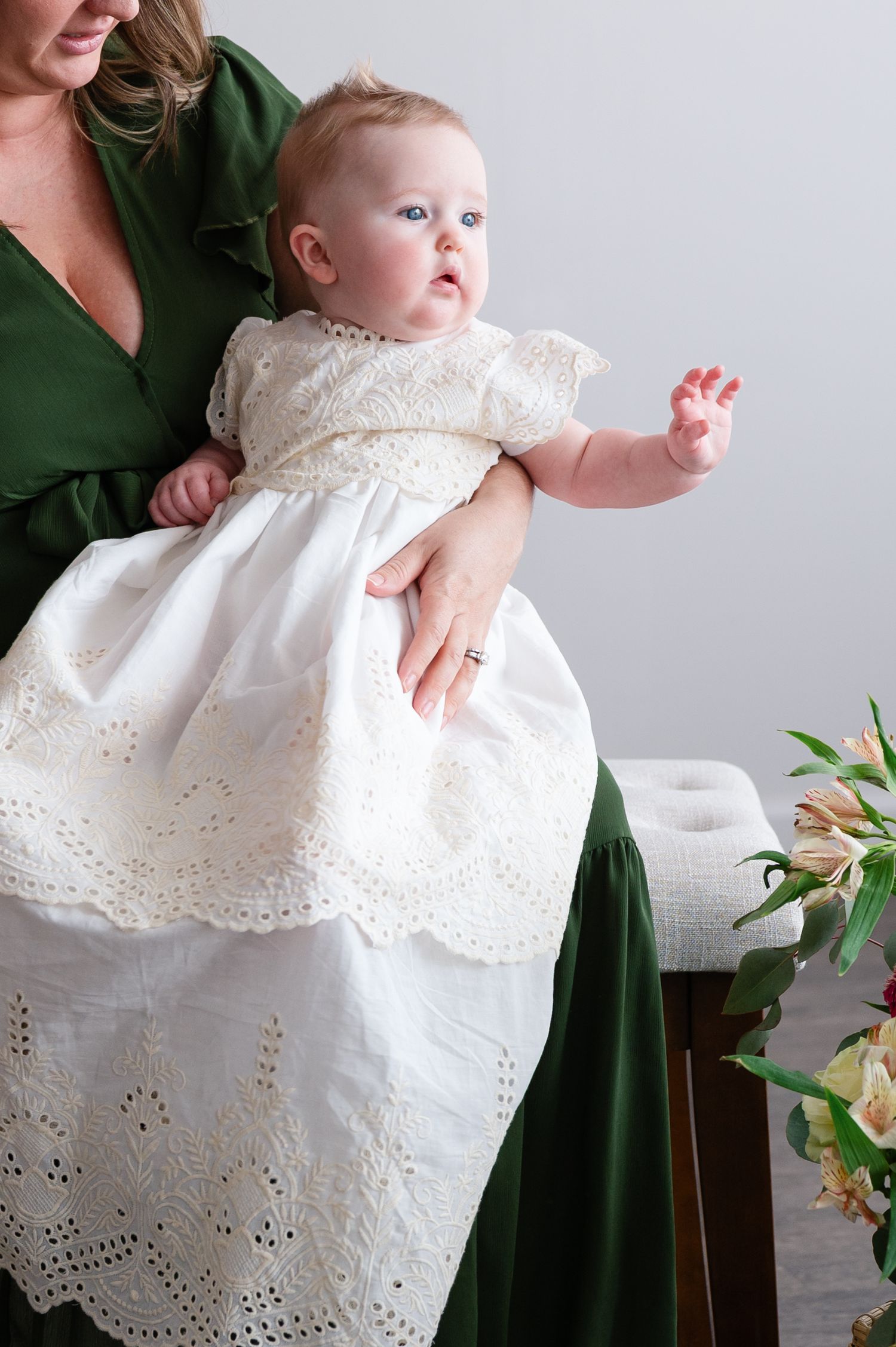 An infant wearing an ornate white lace christening gown sits on someone's lap in a green dress.