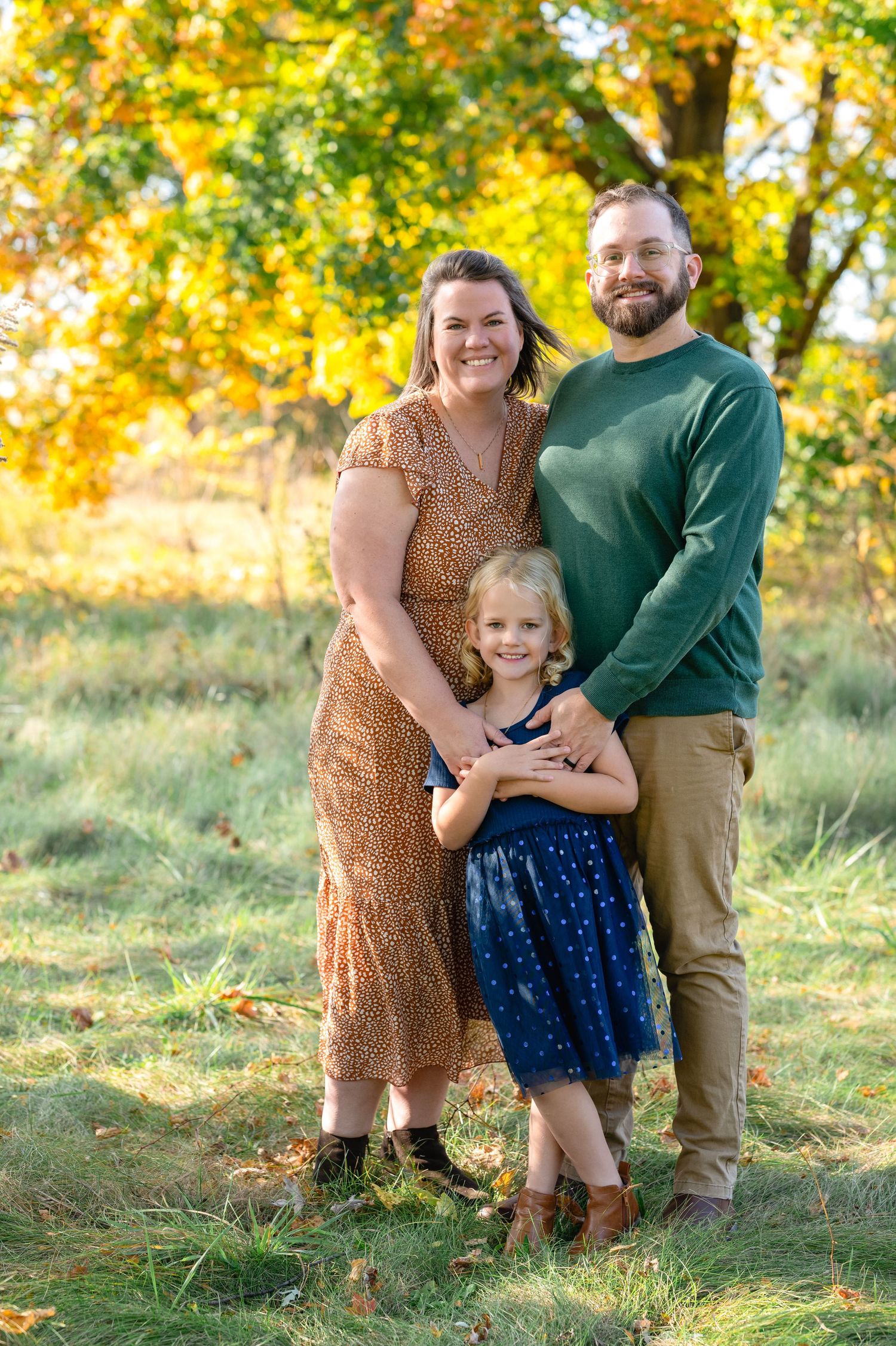 A family poses together in a field during golden hour with autumn trees in the background.