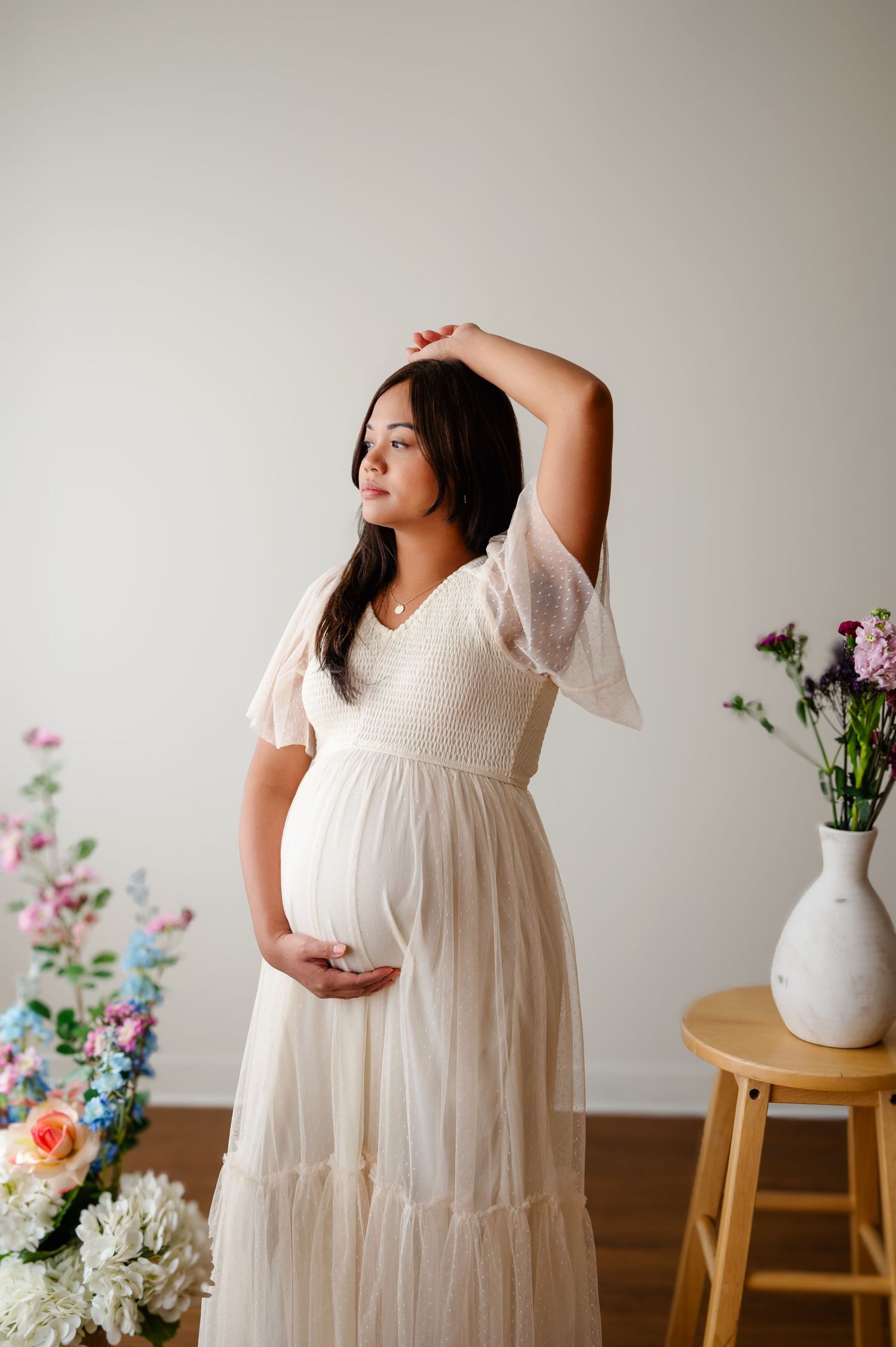 Maternity photo series showing a woman in a flowing white dress posing next to floral arrangements in a minimalist setting.
