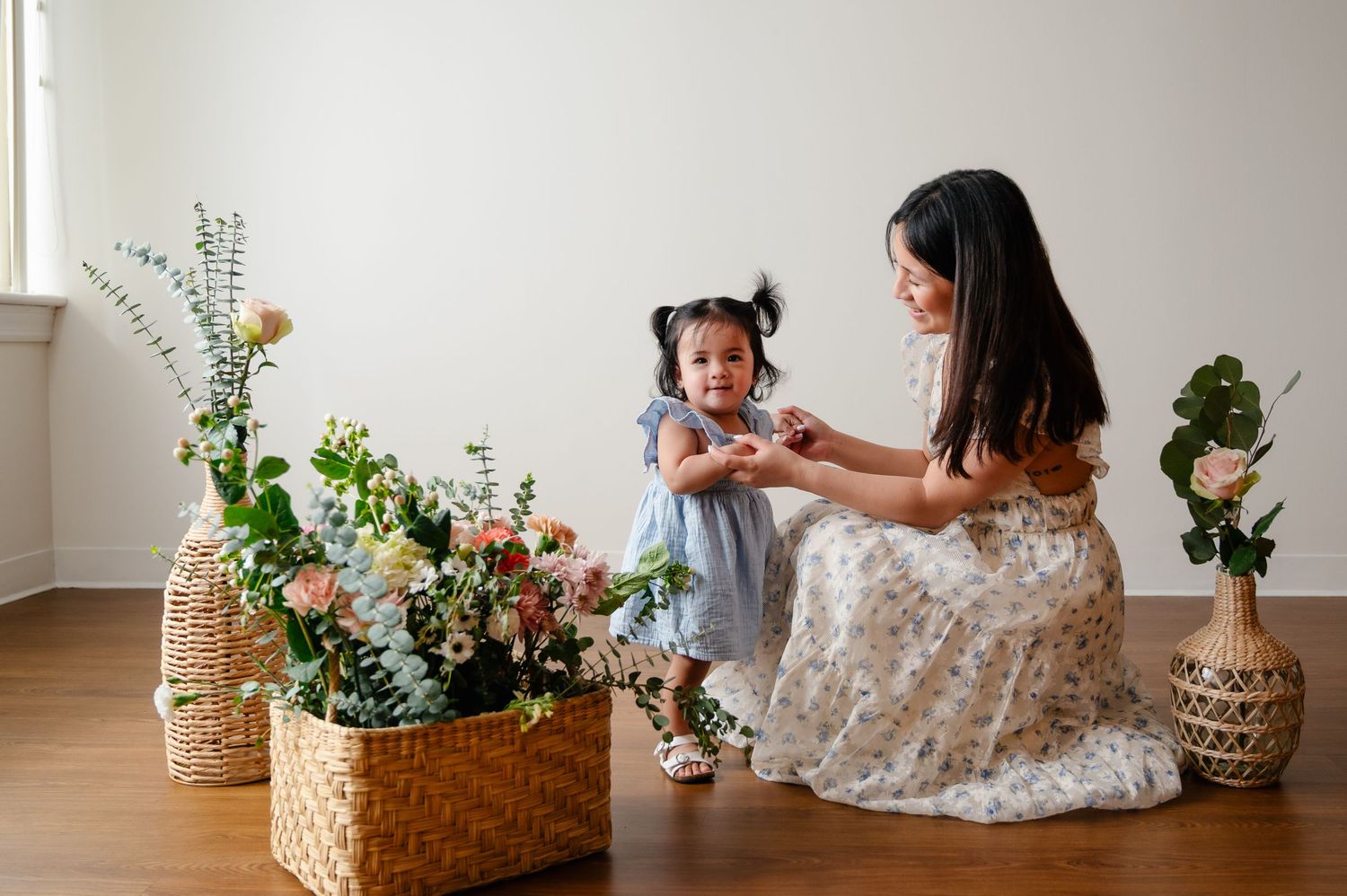 A mother and daughter share a tender moment beside woven baskets filled with fresh spring flowers and greenery.