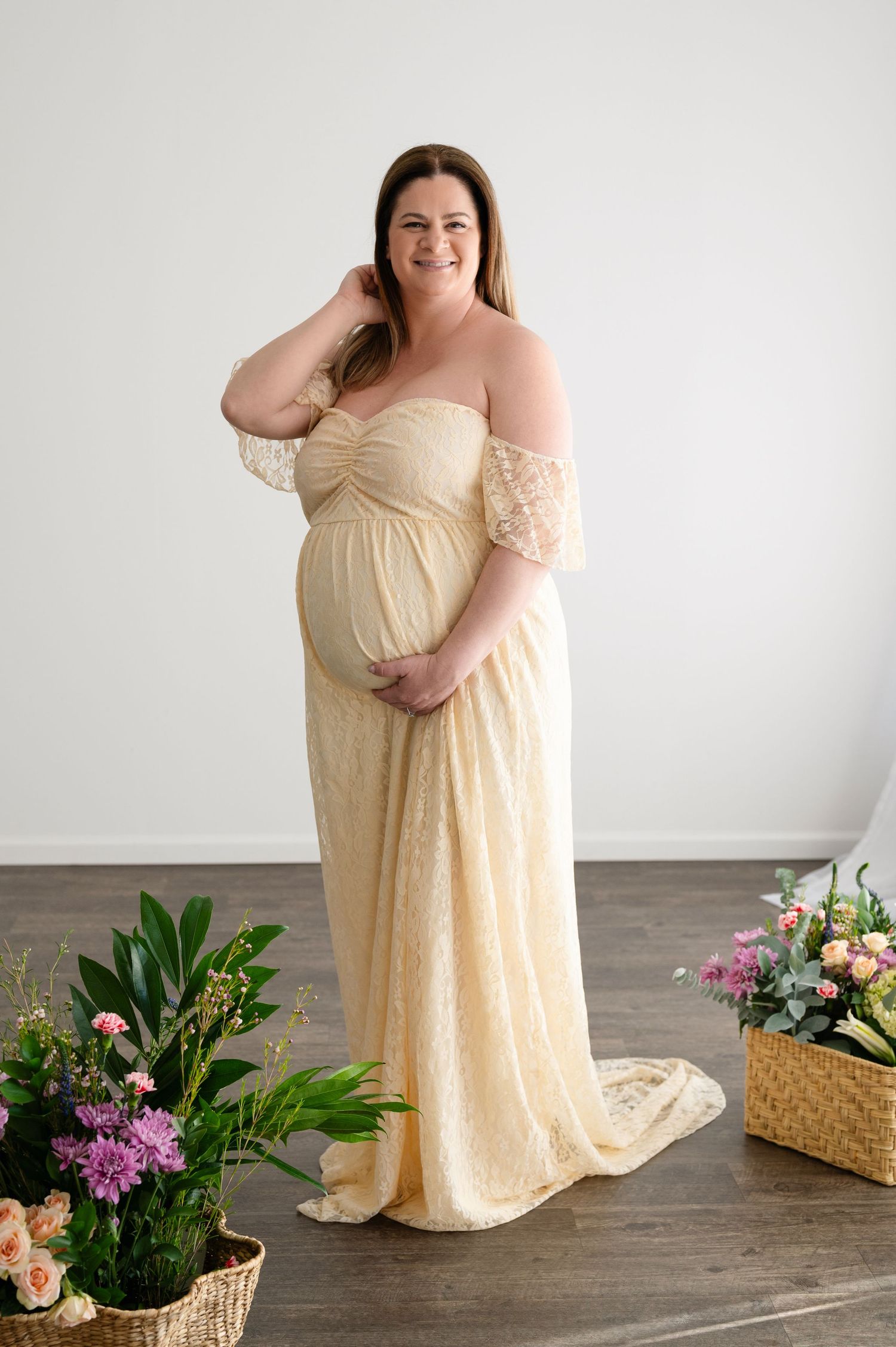 A maternity portrait featuring a woman in a flowing cream-colored lace gown surrounded by flowers.