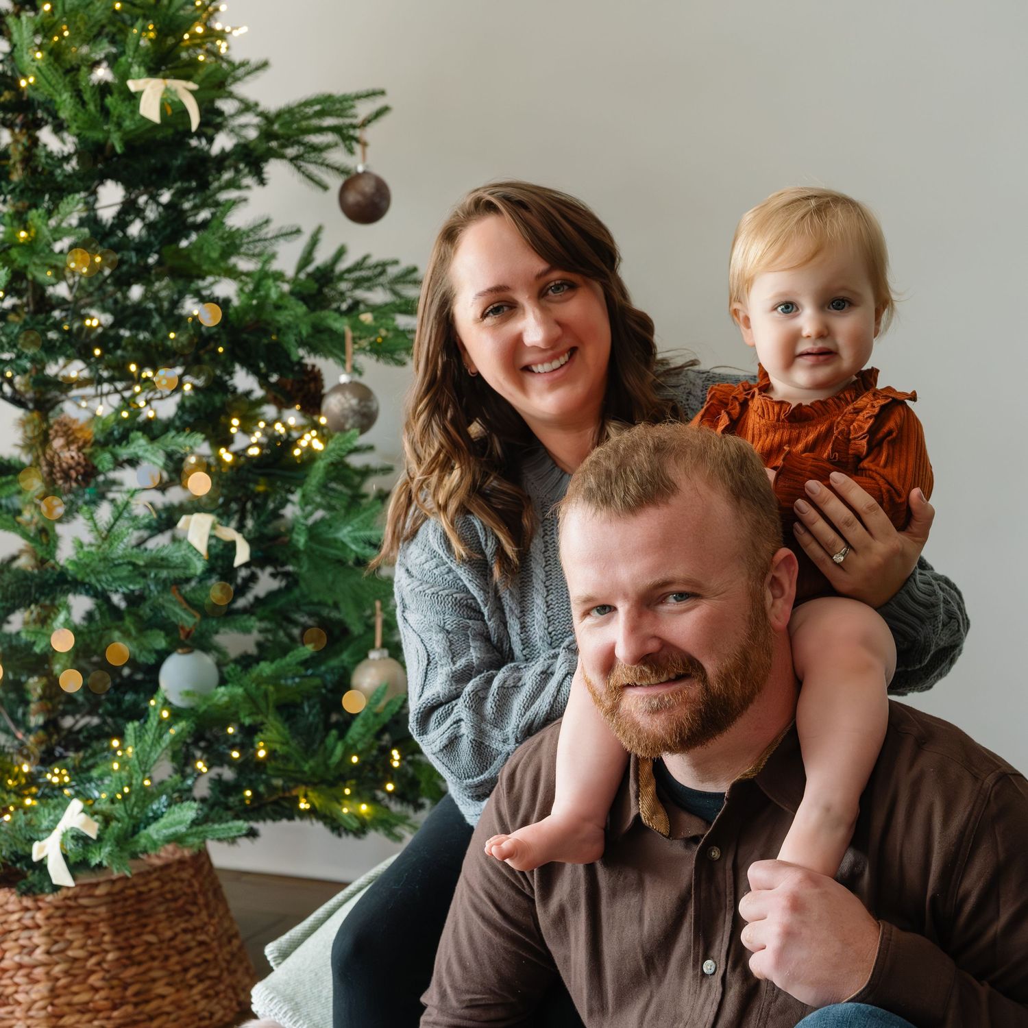 Parents share playful moment with child near Christmas tree during indoor family photoshoot.
