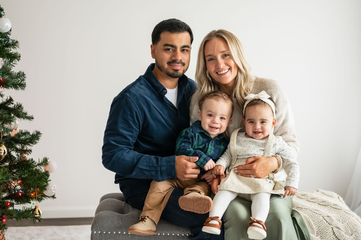 Family gathering on white couch with Christmas tree visible in background during holiday photoshoot.