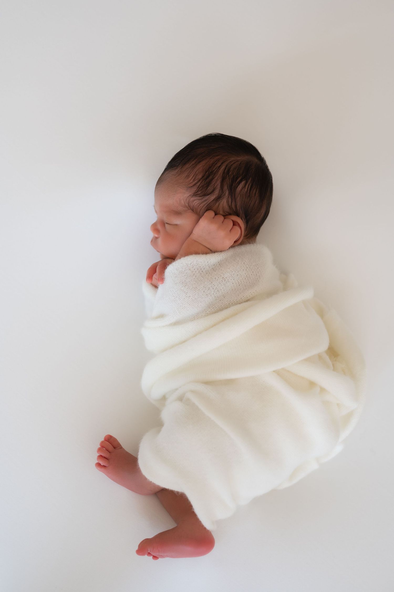 Newborn wrapped in white blanket sleeping peacefully against white backdrop.