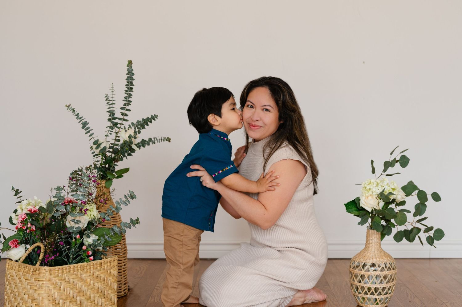 Indoor maternity photoshoot with greenery and floral decorations against white wall.